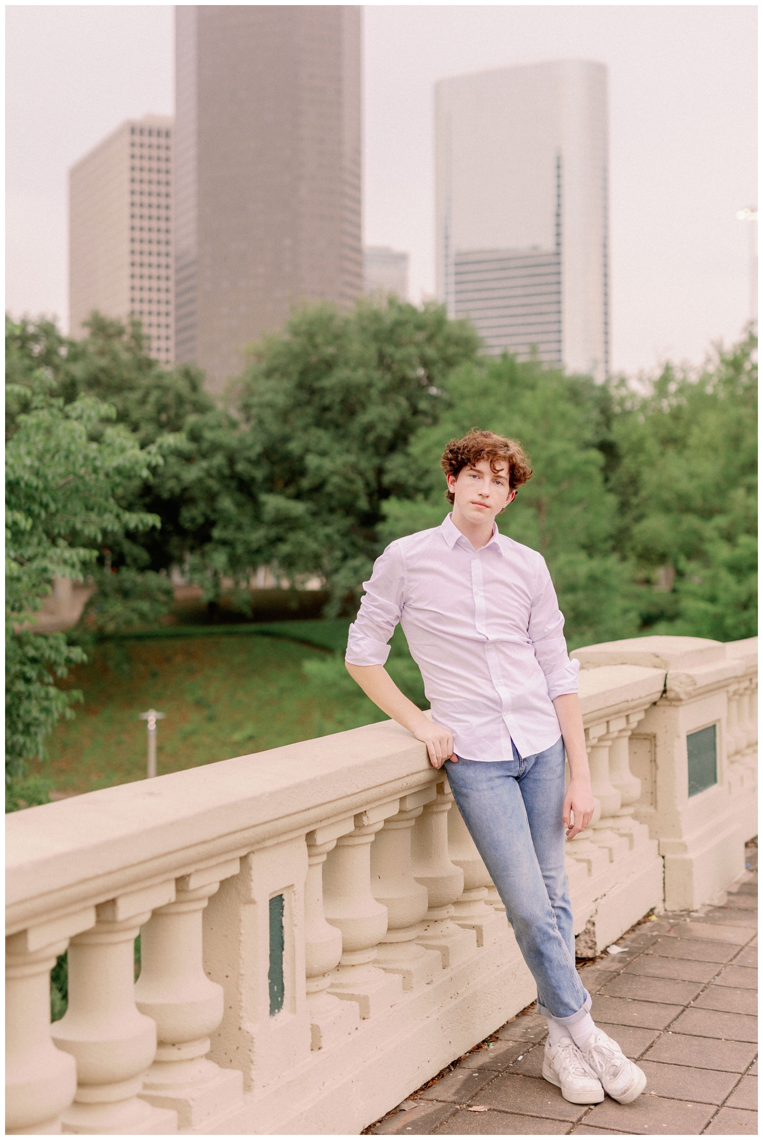 senior boy in white shirt and jeans leaning against bridge in downtown Houston in front of skyline