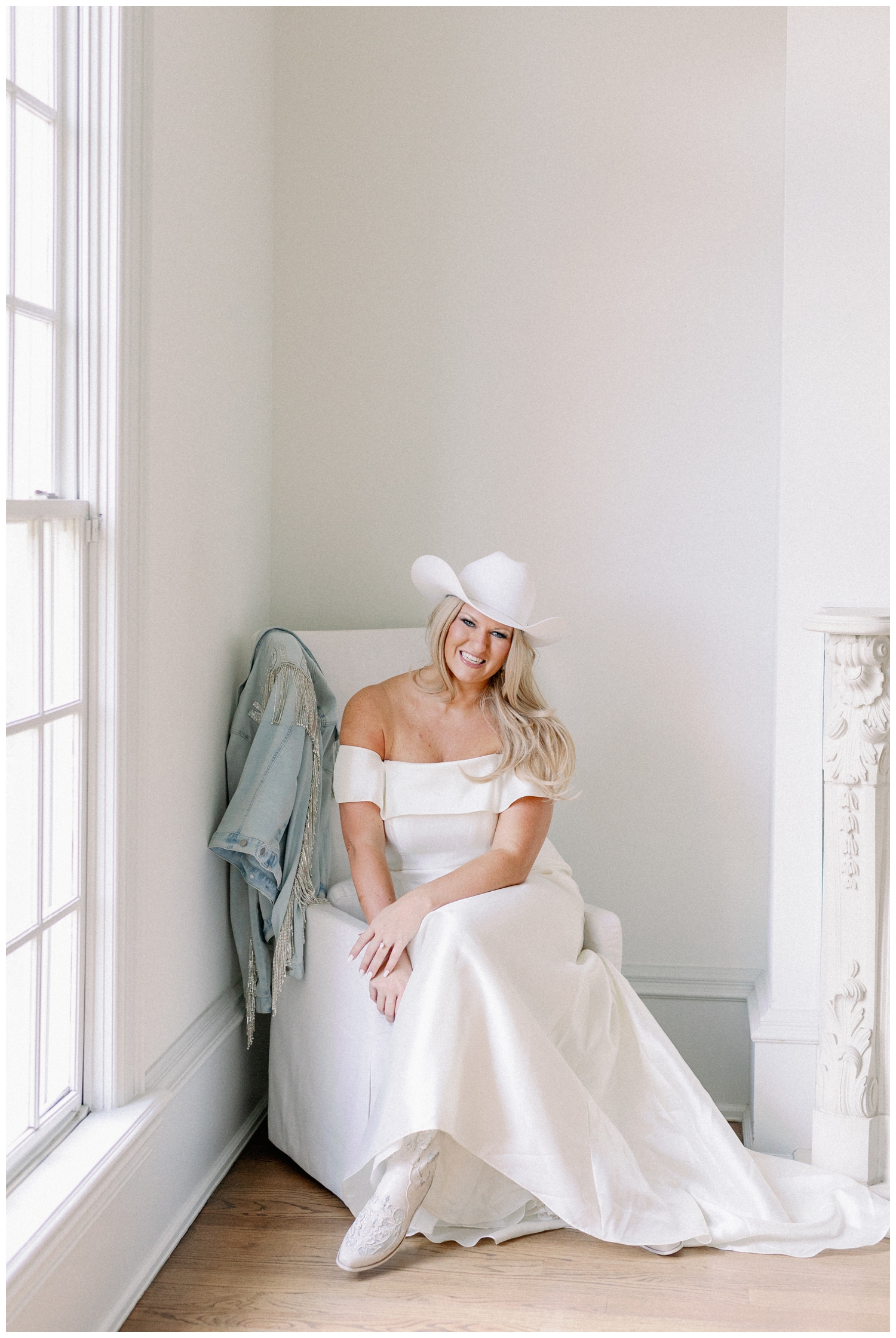 bride sitting on white chair in wedding dress with monogrammed jacket, white boots and white cowboy hat