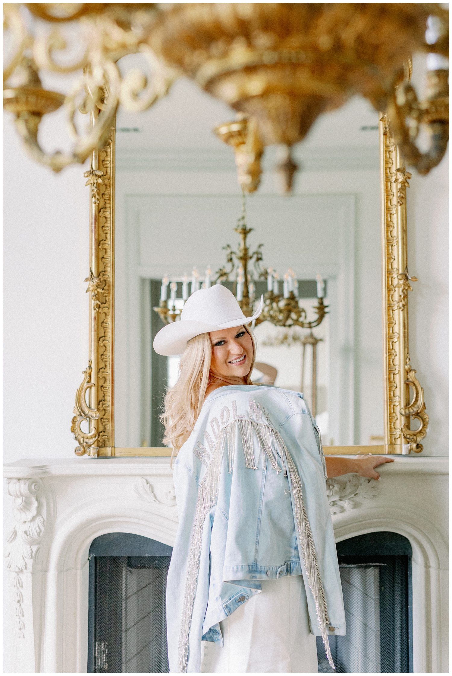 bride holding monogrammed denim jacket with white cowboy hat in front of gold mirror