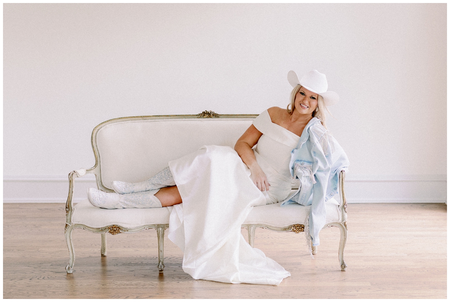 bride in wedding gown sitting on couch with white boots, white cowboy hat and denim jacket