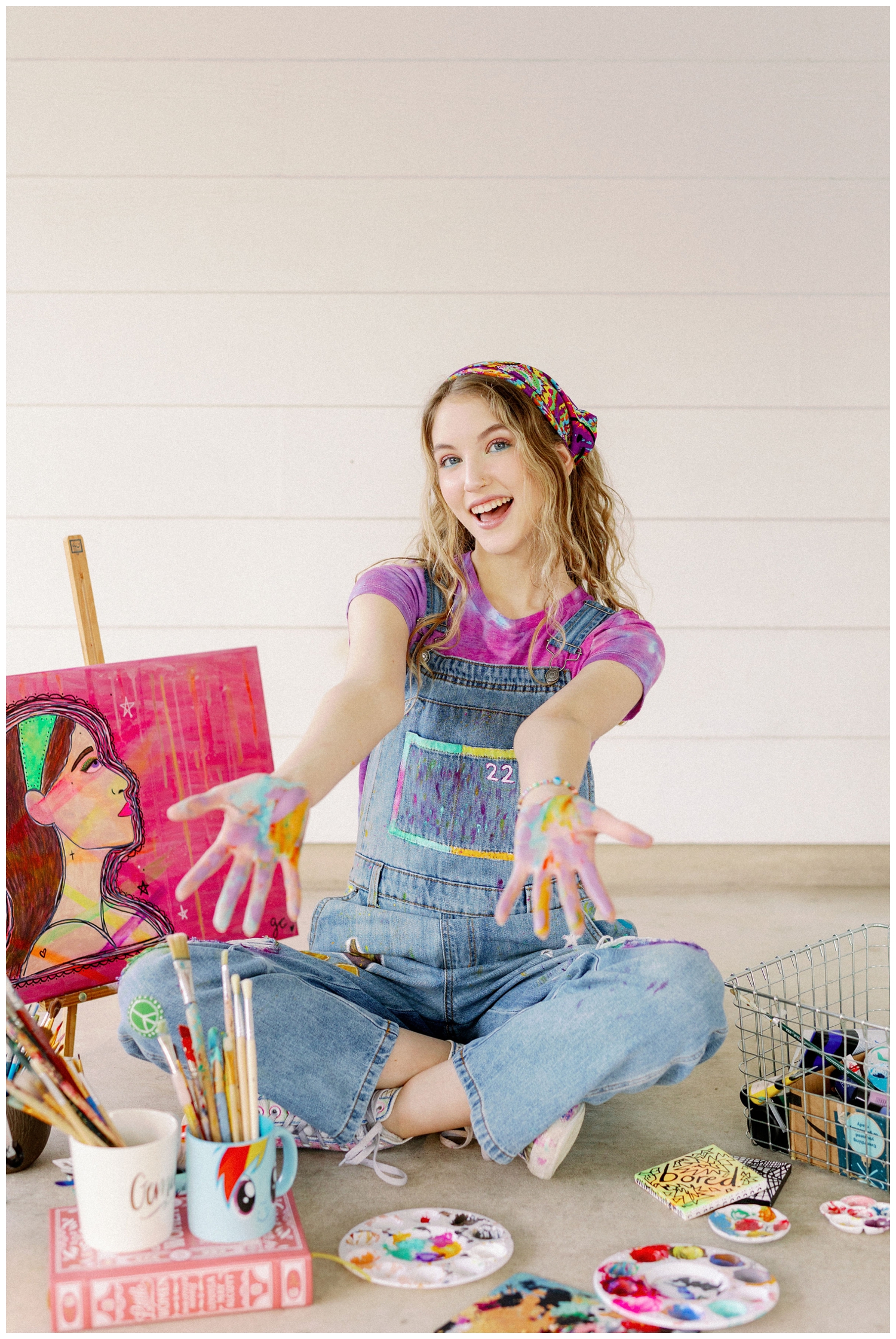 girl seated in overalls holding out painted hands for colorful Houston senior photos