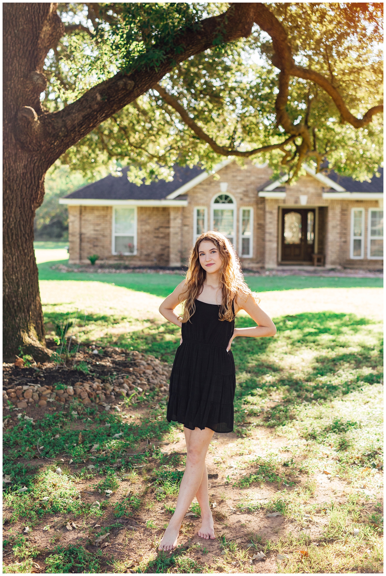 senior girl in black sundress with hands on hip standing under tree in front of her home