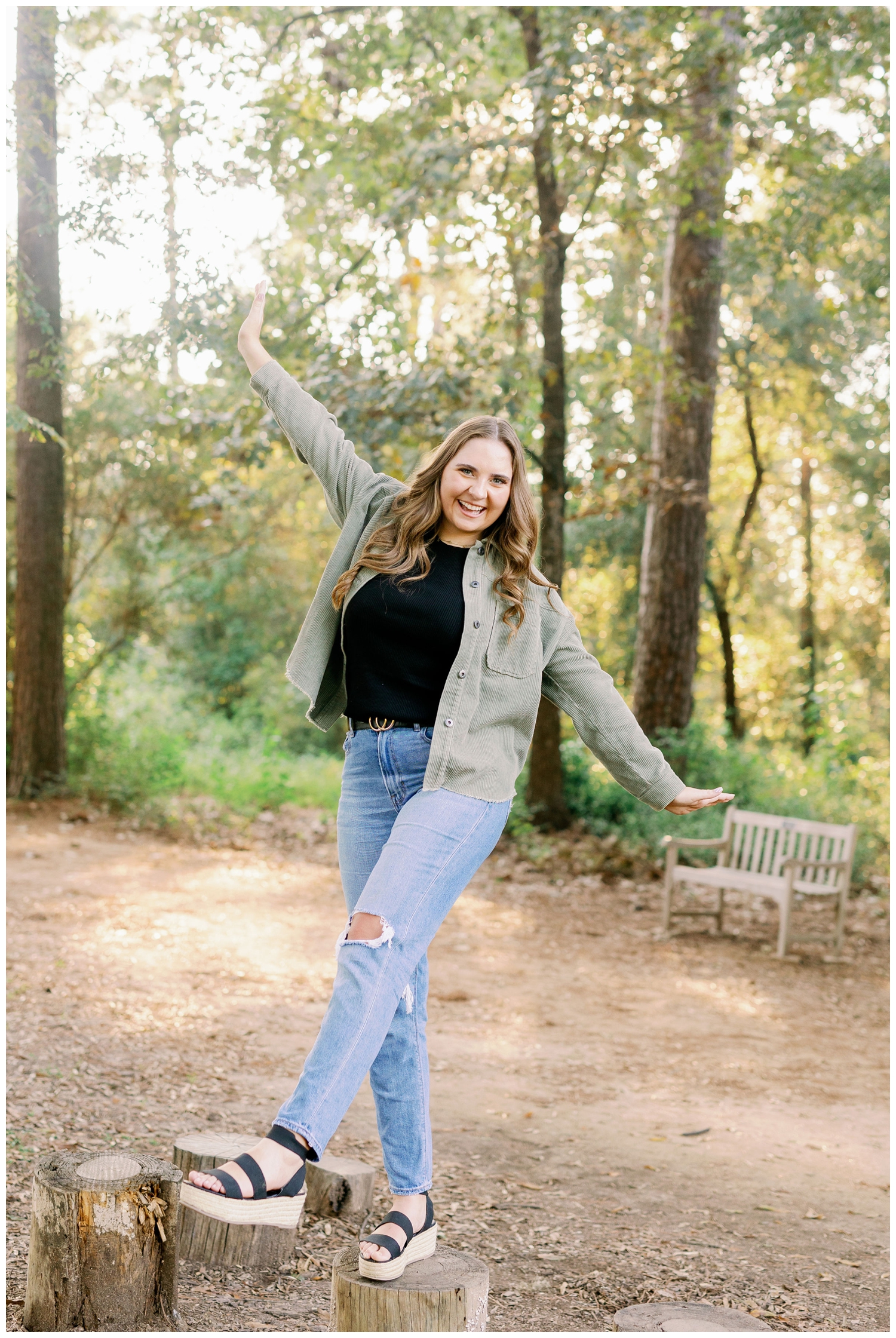 girl in jeans and green denim jacket standing on a stump by trees for senior pictures Houston Arboretum