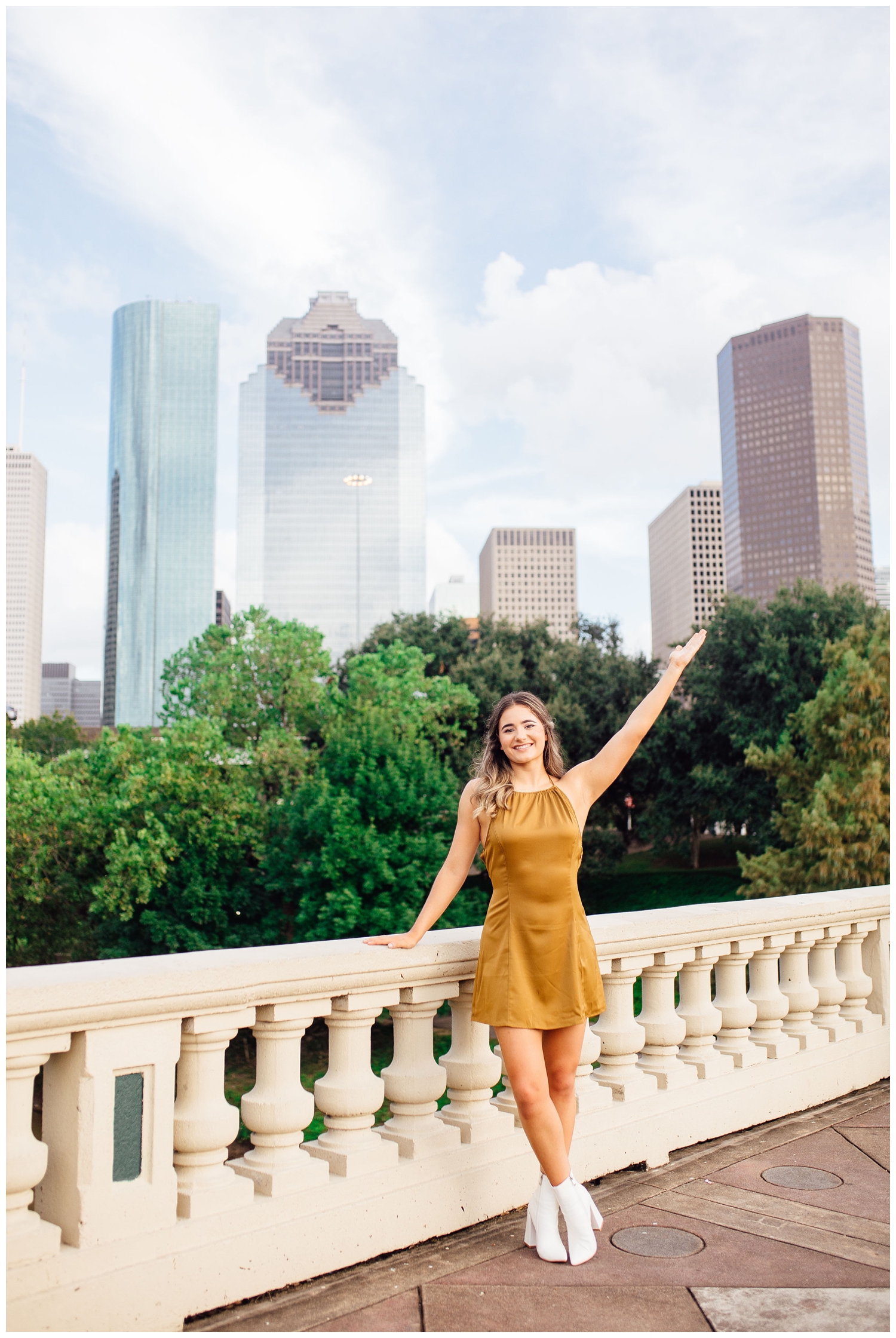 girl in white boots and gold dress with arm in air standing in front of Houston skyline