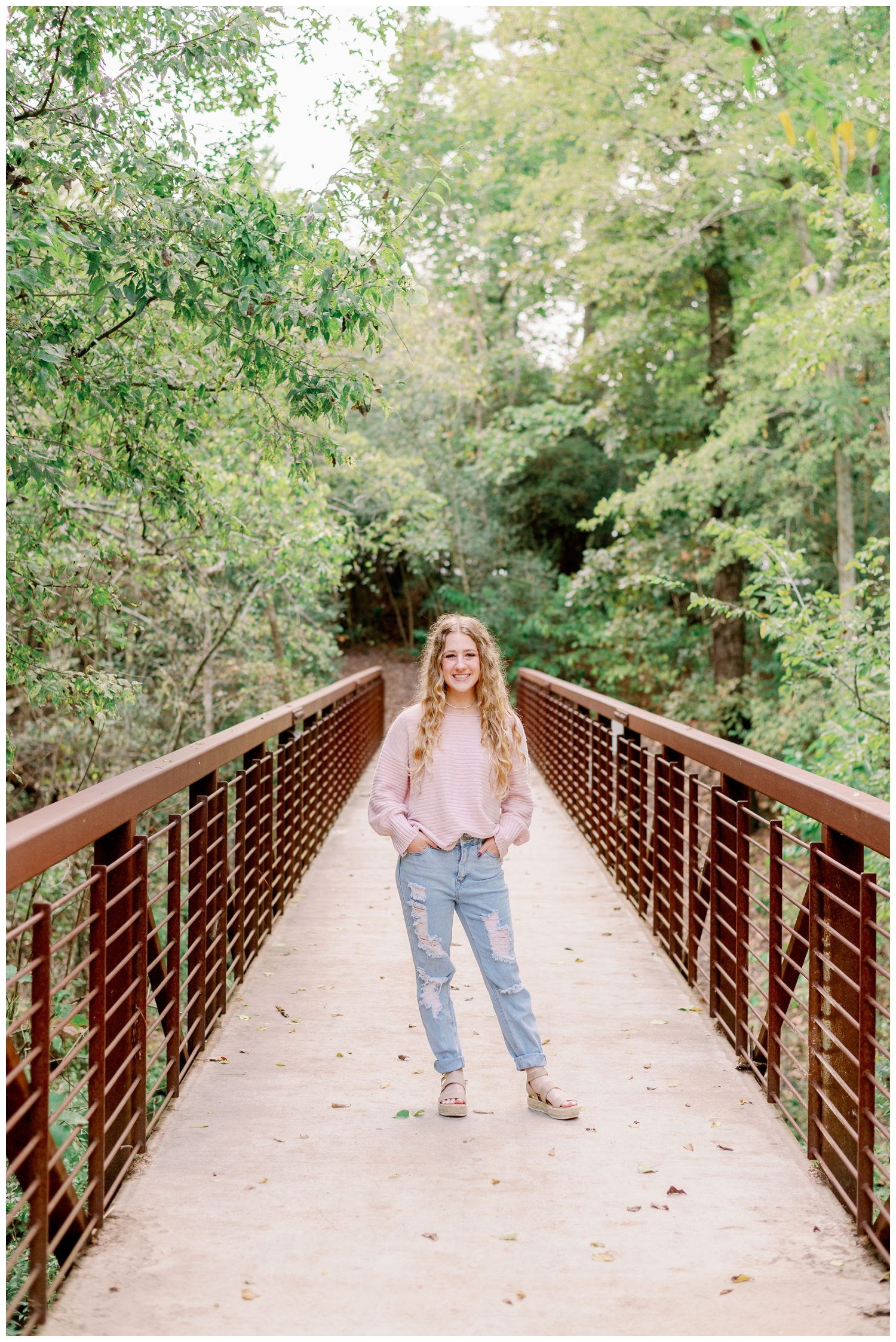high school senior girl in light blue jeans and pink shirt standing on bridge with hands in pocket at Houston Arboretum