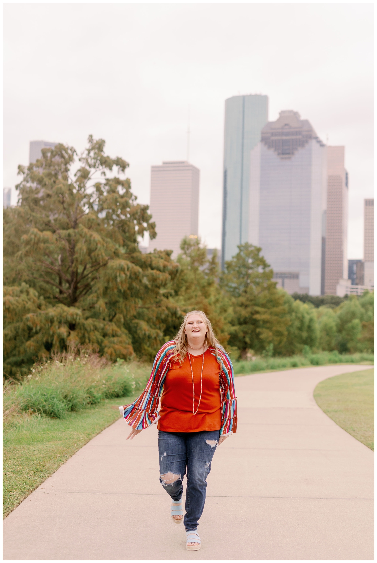girl in jeans and colorful shirt walking on pathway with Houston skyline in background