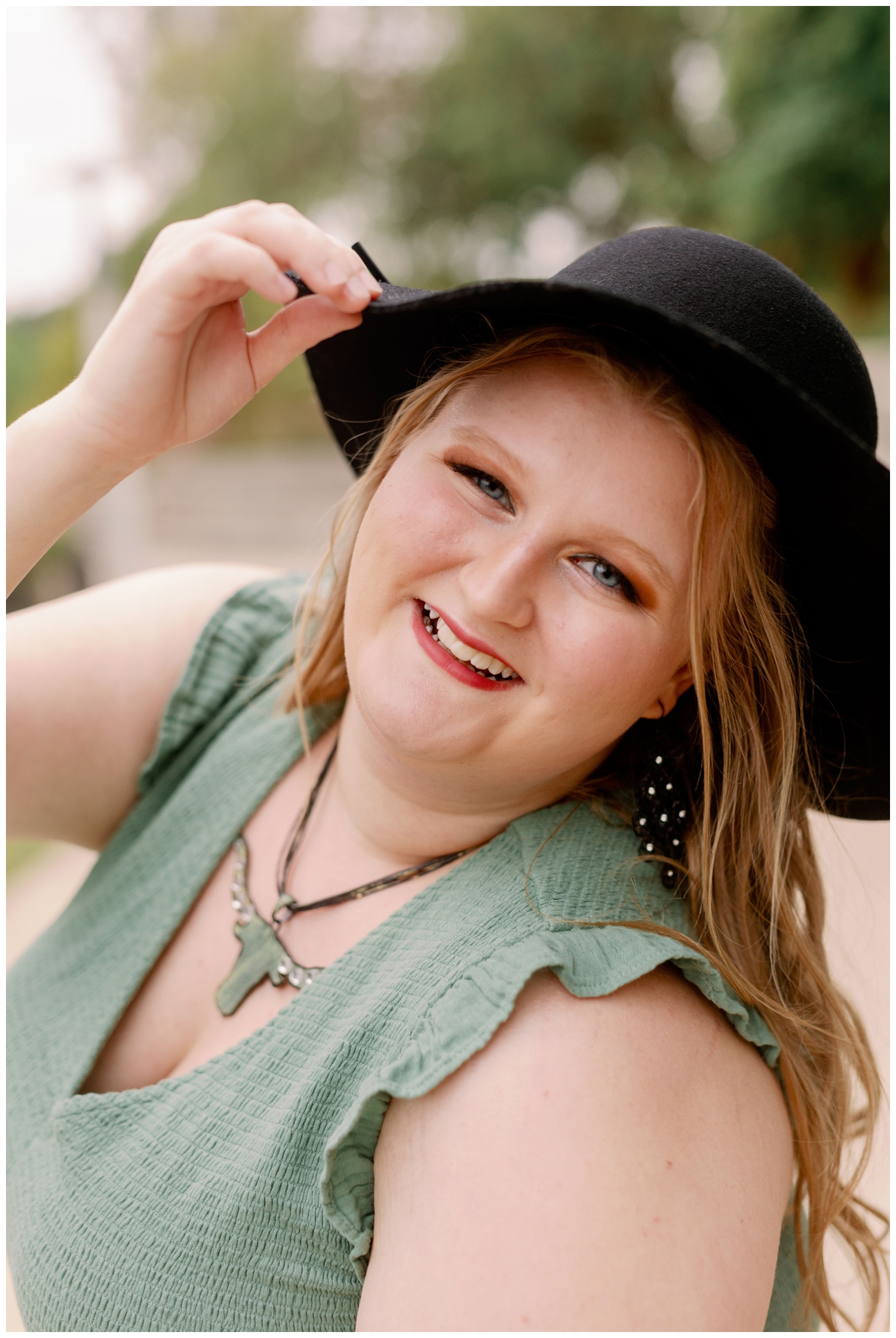 girl in sage dress with black hat leaning toward camera at for senior photos Eleanor Tinsley Park in Houston