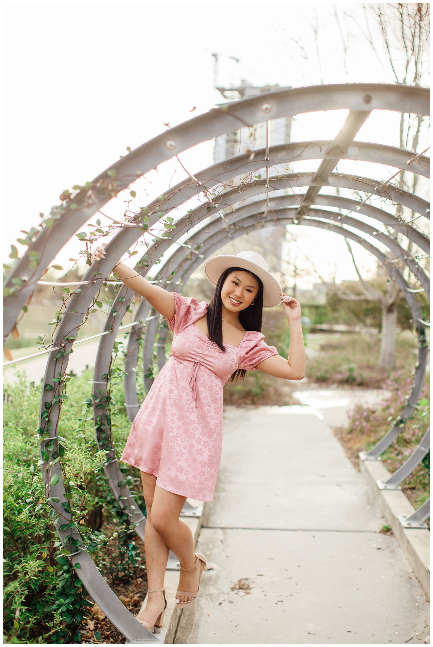 high school senior in pink dress with white hat hanging on garden arches in Houston