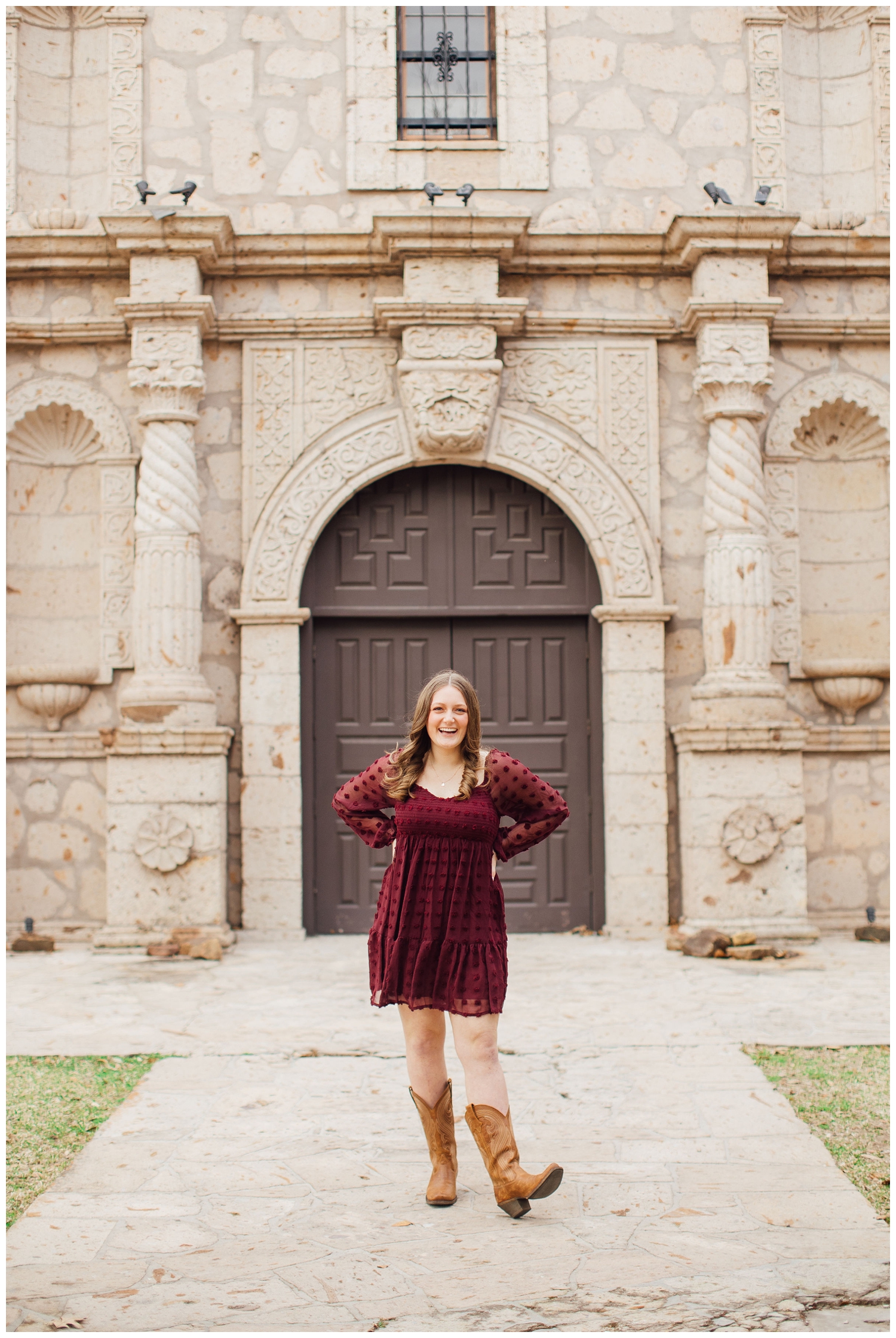 high school senior girl in maroon dress standing with hands on hip in front of rustic building