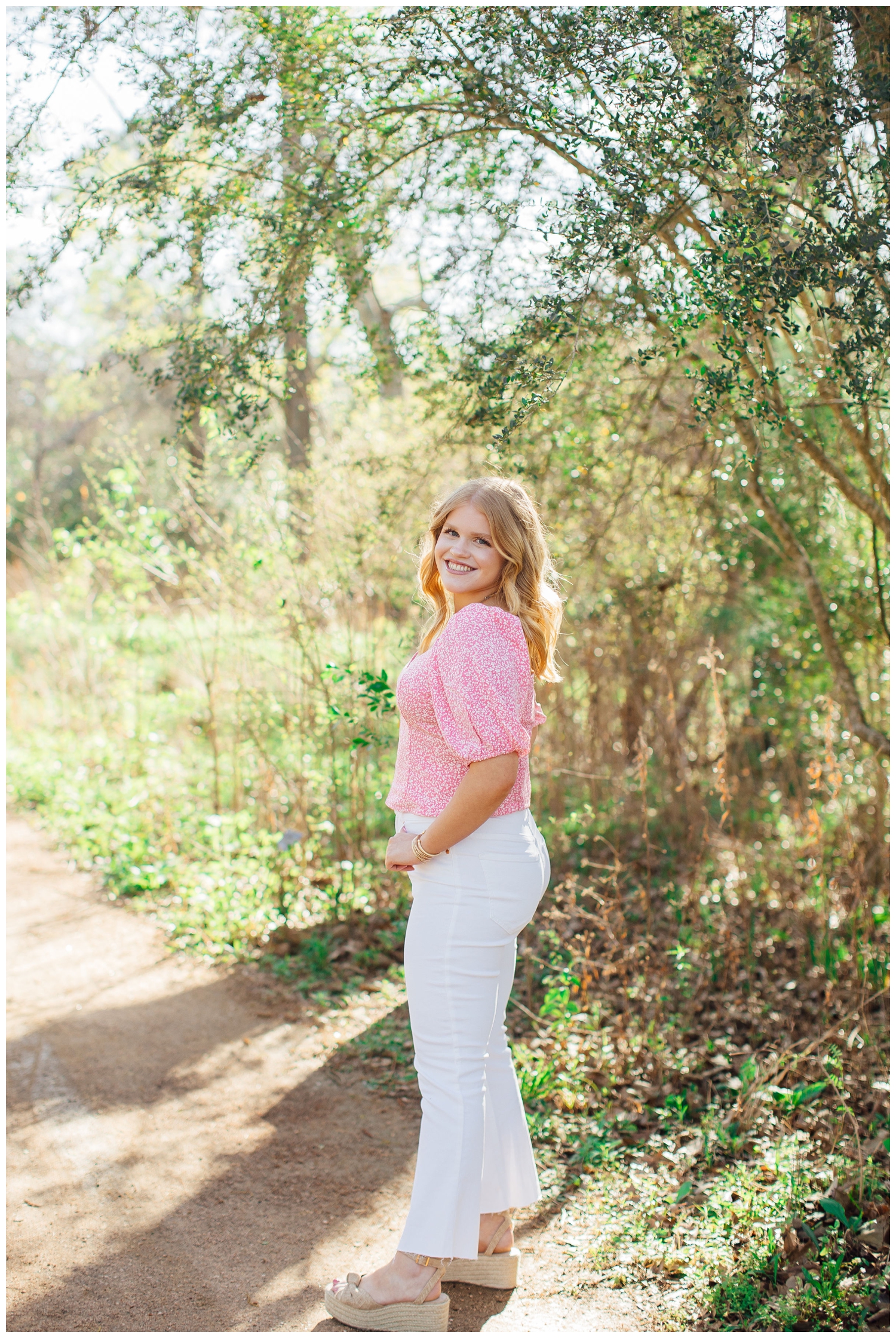 senior girl in pink shirt and white pants looking over her shoulder on pathway at Houston Arboretum