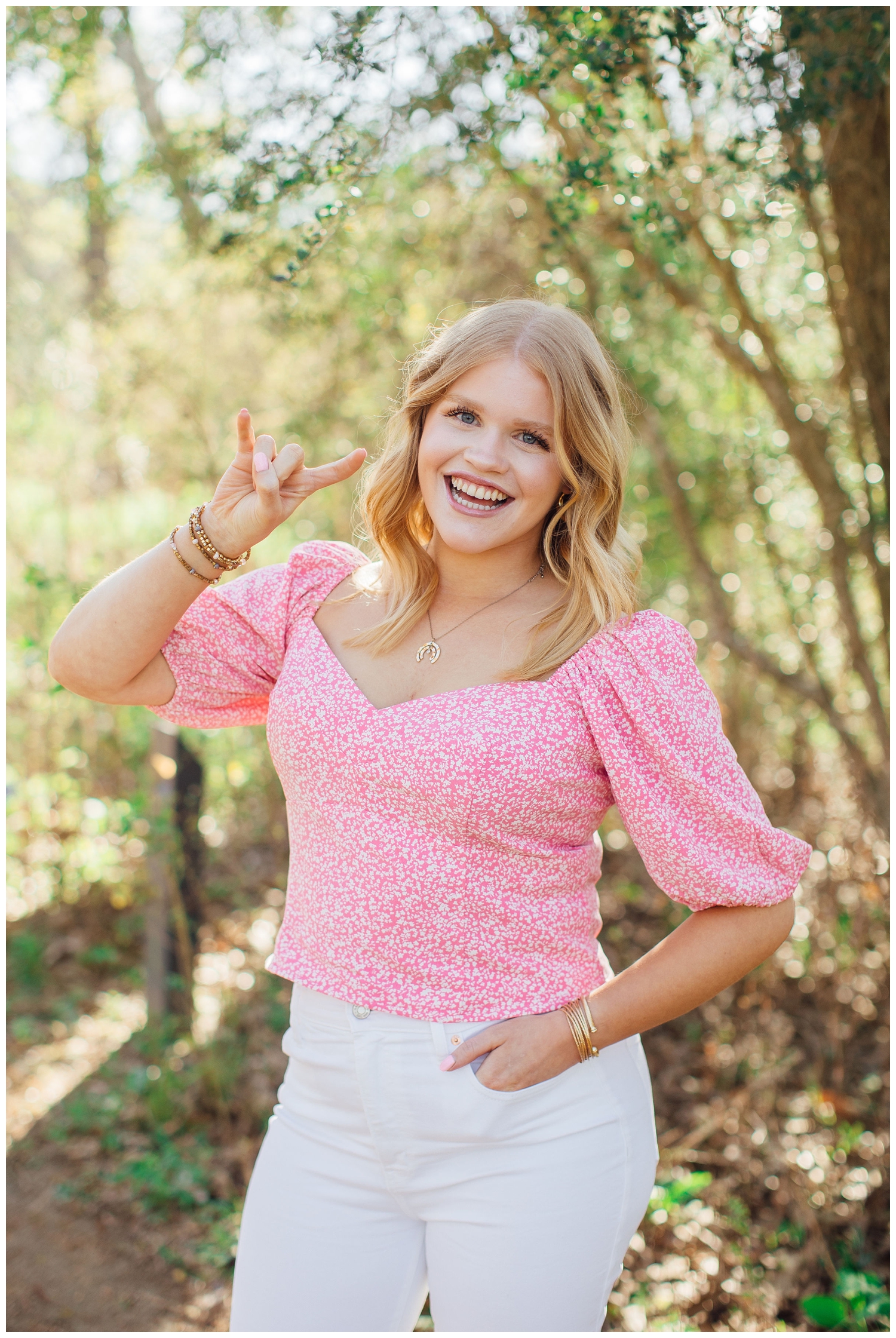high school senior girl in white pants pink shirt in front of trees with college hand sign