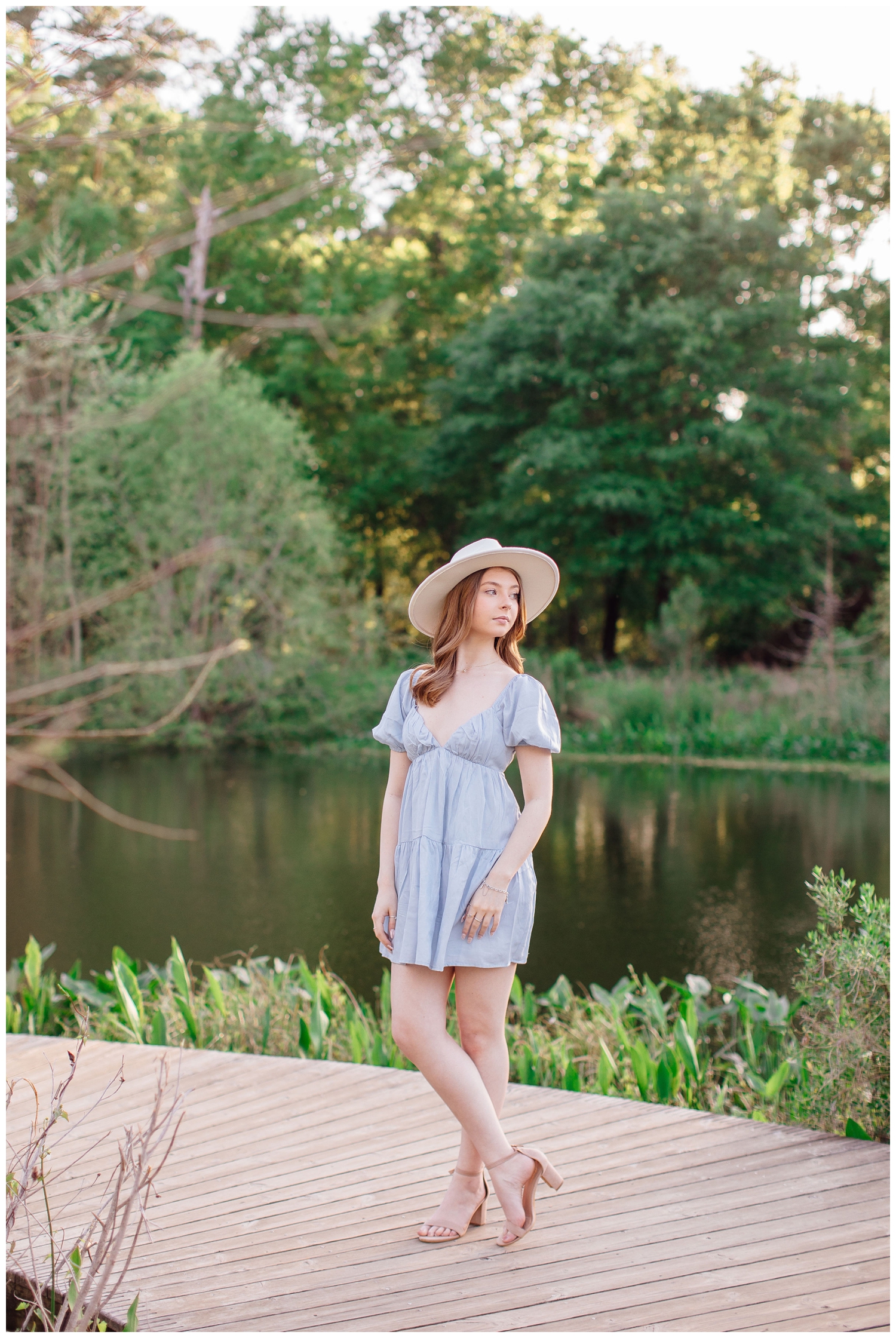 Houston senior portrait with girl standing in blue dress legs crossed white brimmed hat in front of pond