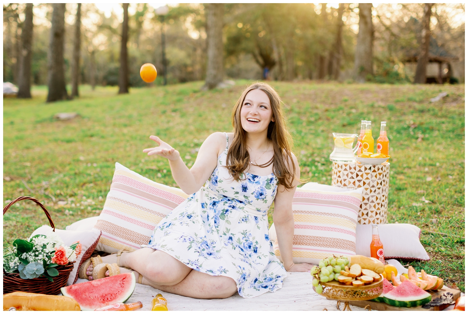 spring picnic senior pictures of girl sitting on blanket in floral dress throwing orange in air