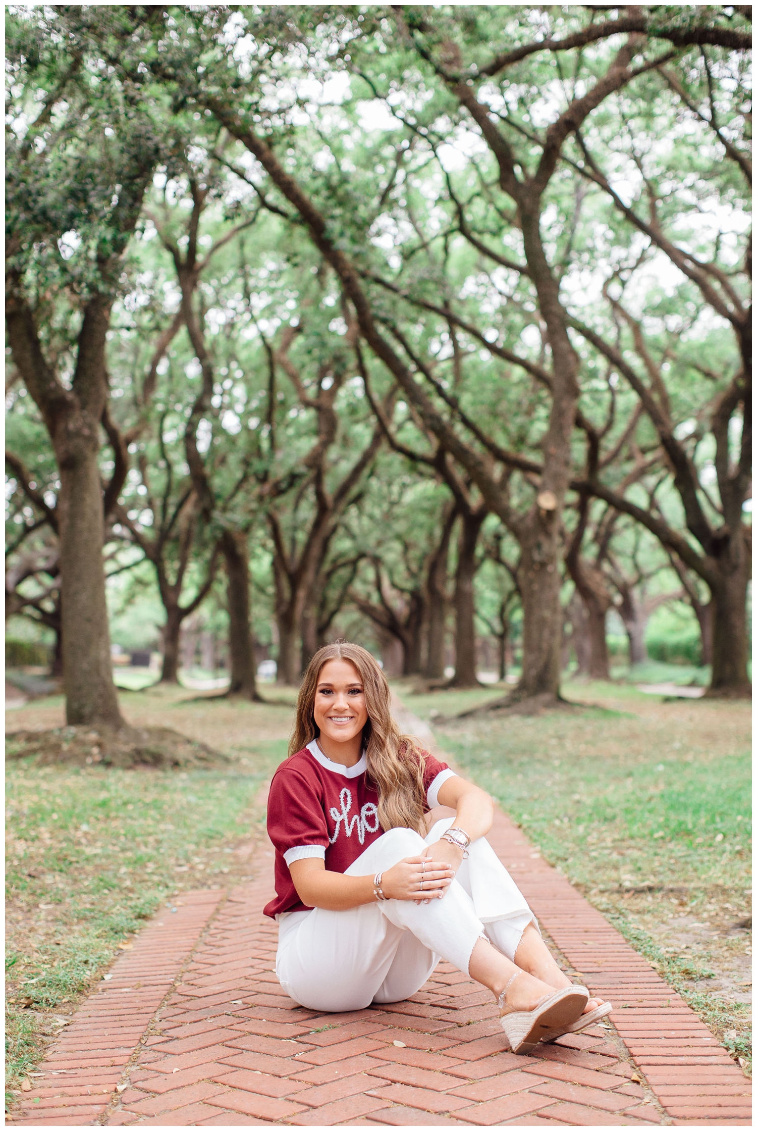 high school senior girl sitting on path between trees at North South Blvd in Houston wearing white pants and maroon shirt