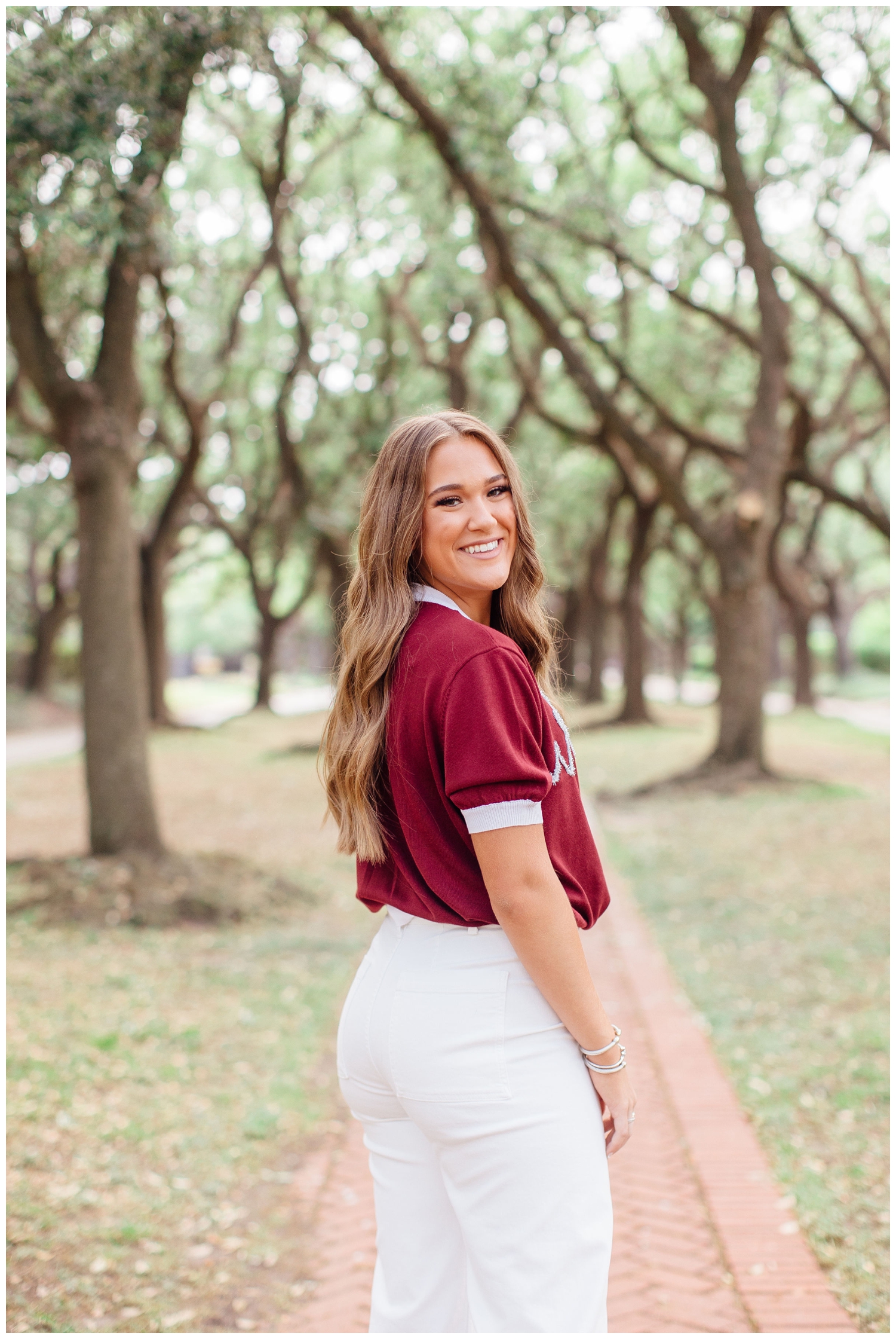 girl in maroon shirt white jeans standing in front of trees smiling