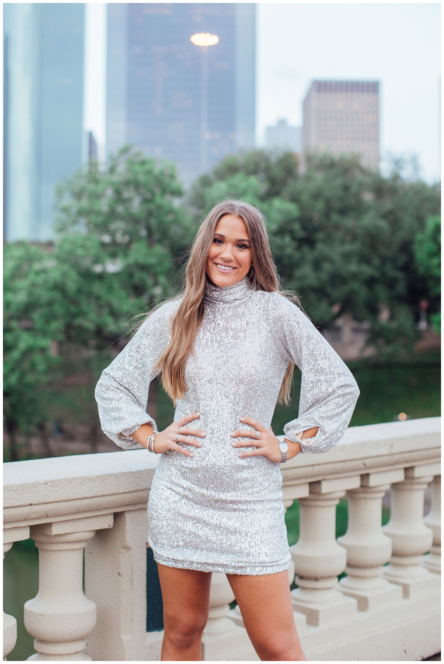 high school senior in silver sequence dress standing in front of Houston skyline with hands on hip