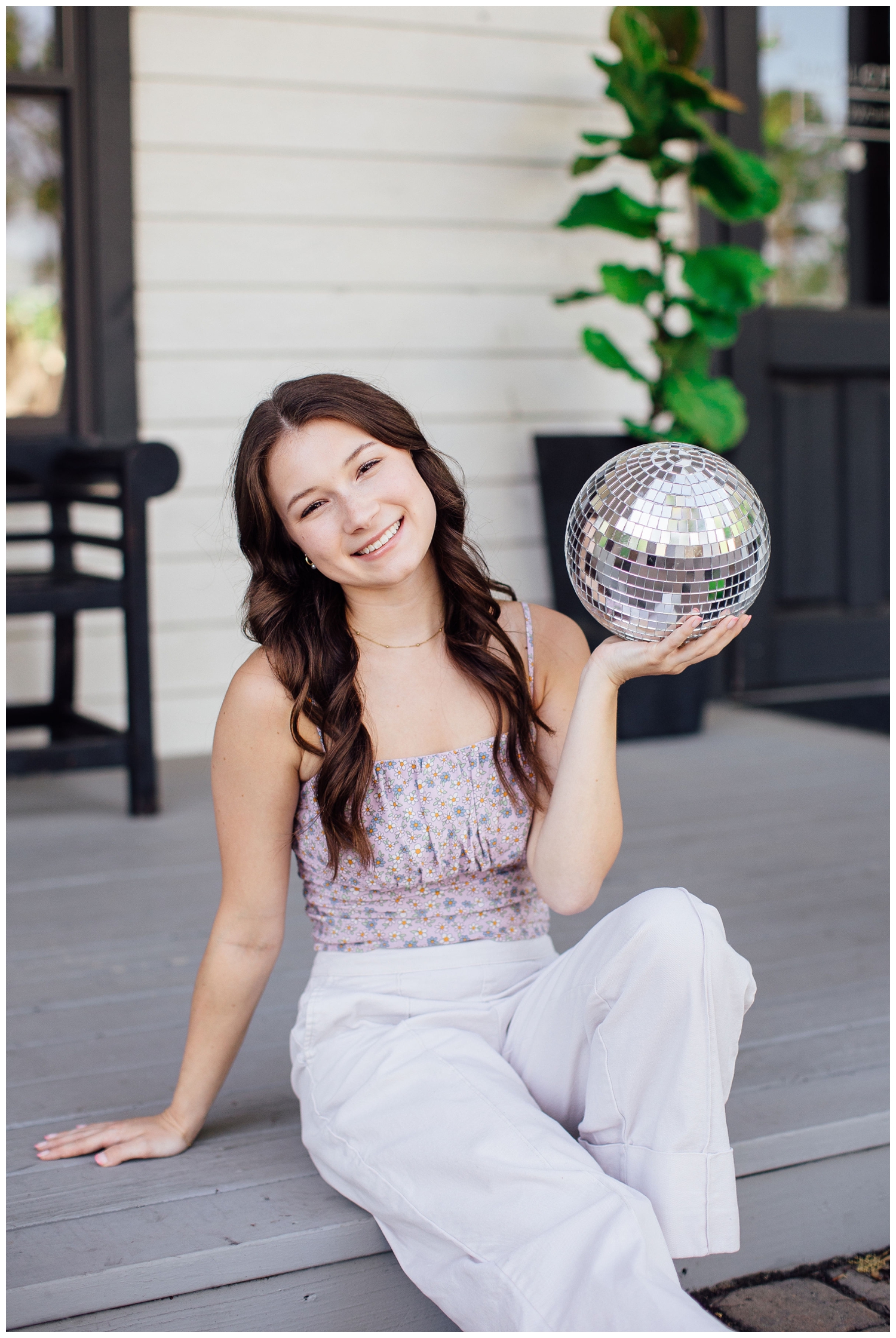 girl in white pants sitting on ground holding silver disco ball for spring senior photos Houston