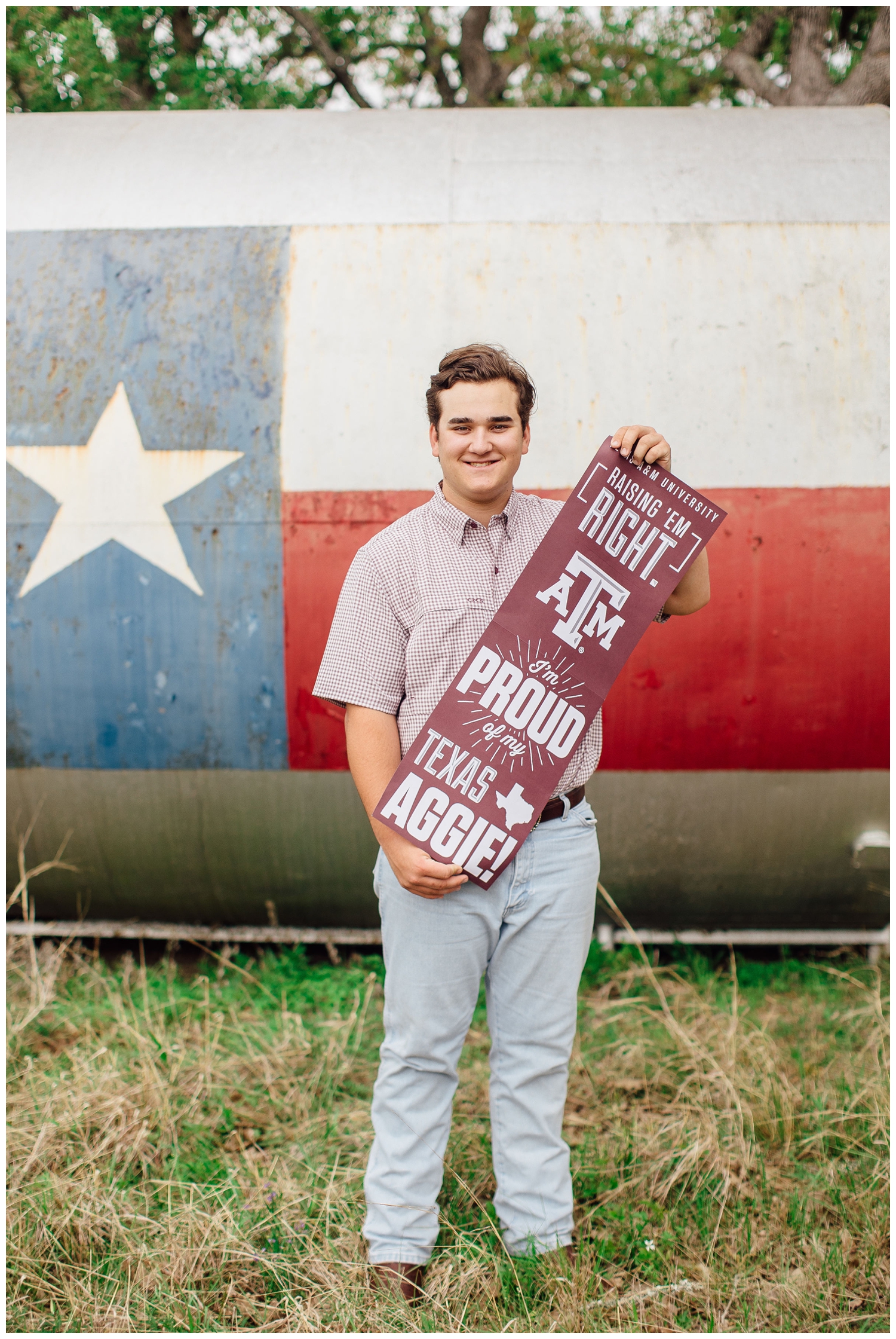 high school senior boy in jeans and plaid shirt holding Texas Aggie sign in front of lone star barrel
