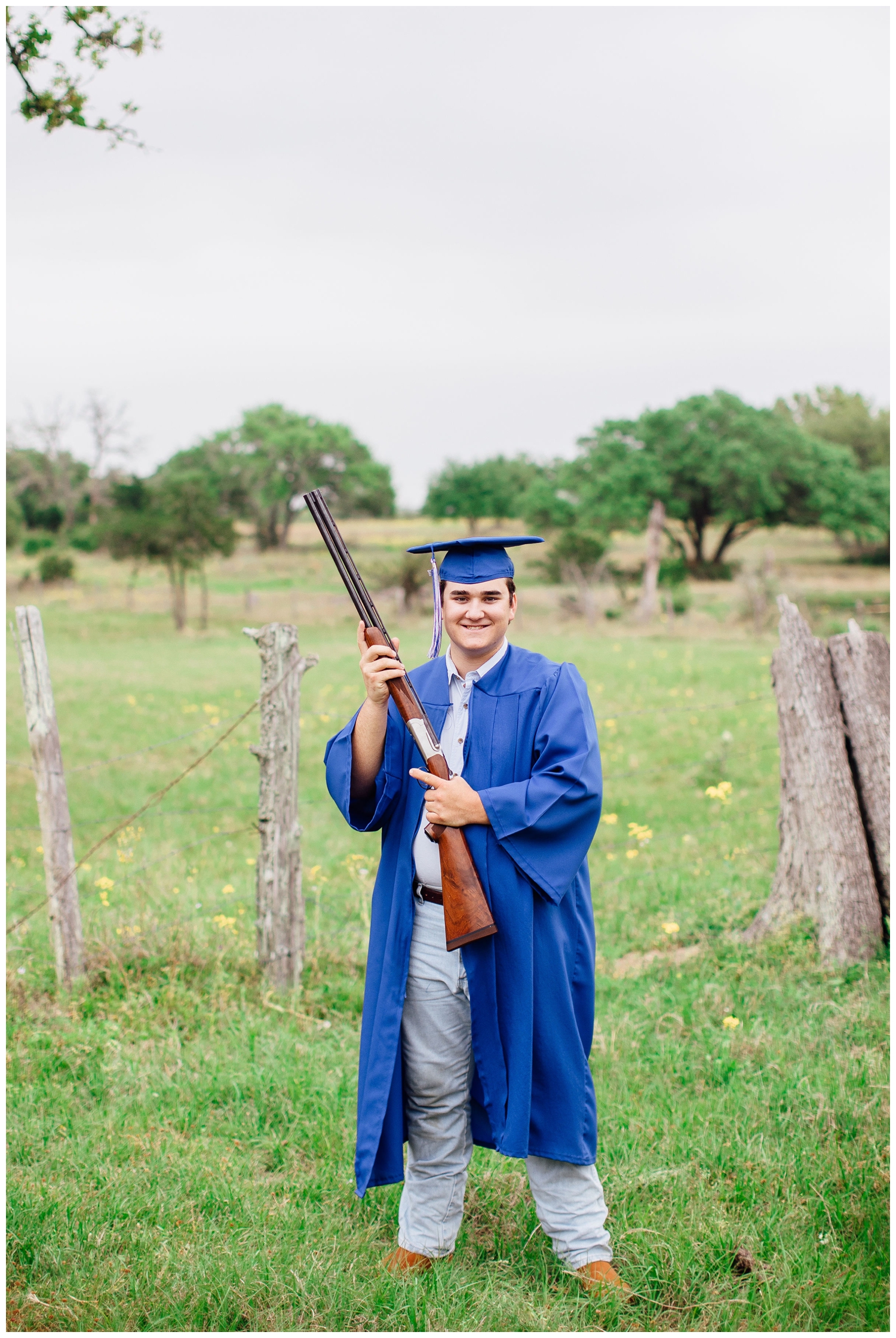 high school guy standing in field wearing blue cap and gown holding gun for senior pictures for guys Houston, Texas
