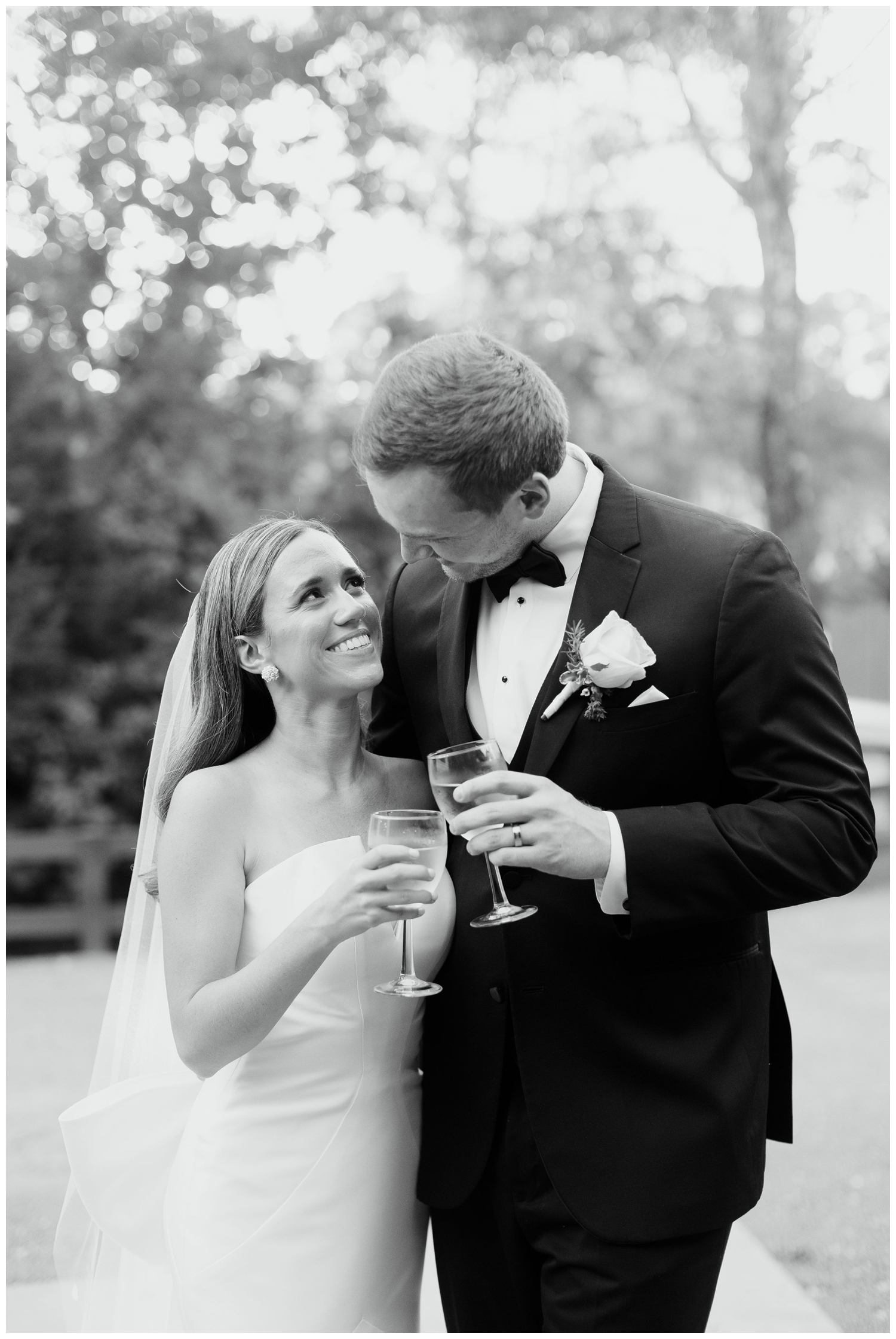 black and white image of bride and groom smiling and holding champagne glasses outdoors