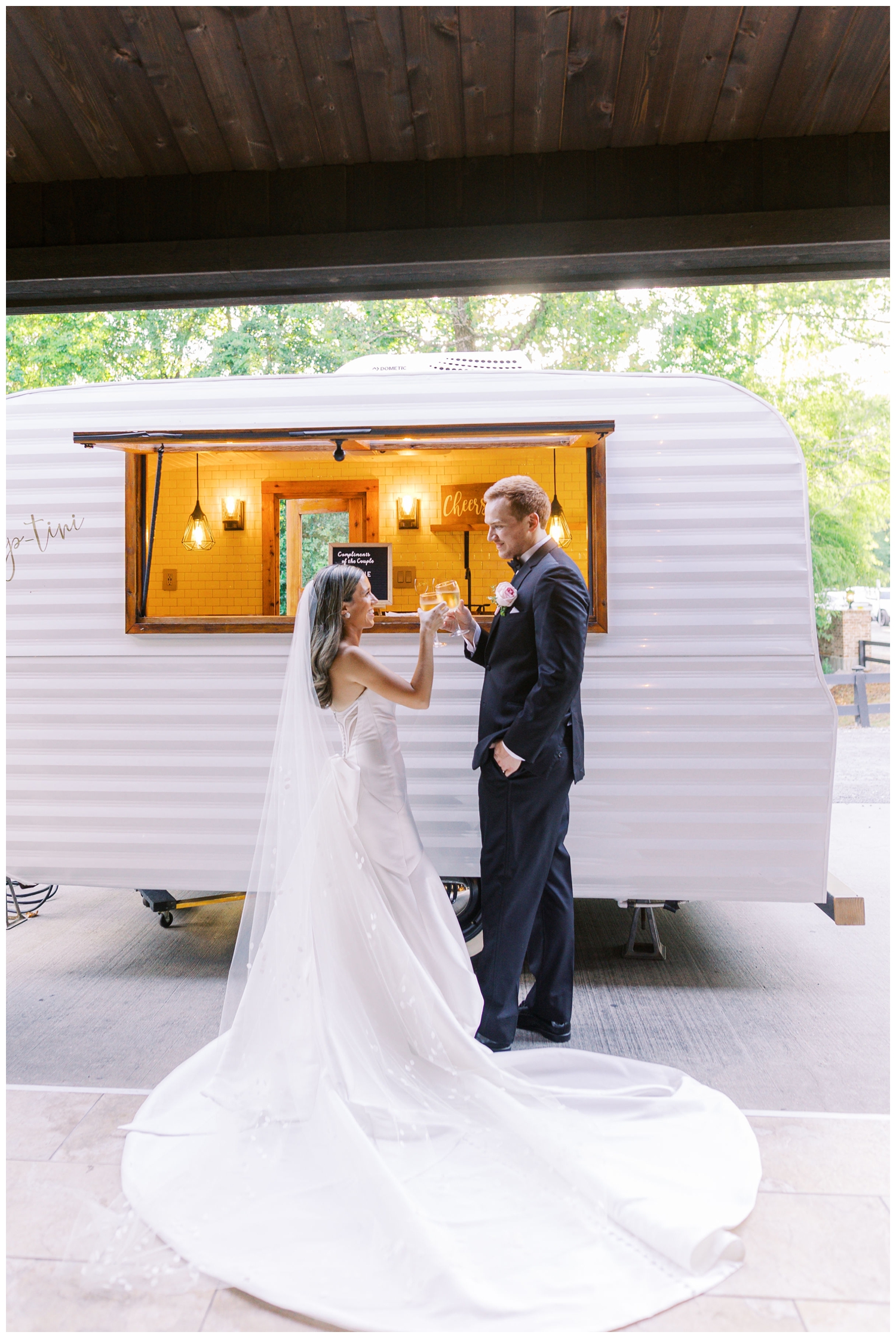 bride and groom standing in front of white mobile bar camper drinking champagne