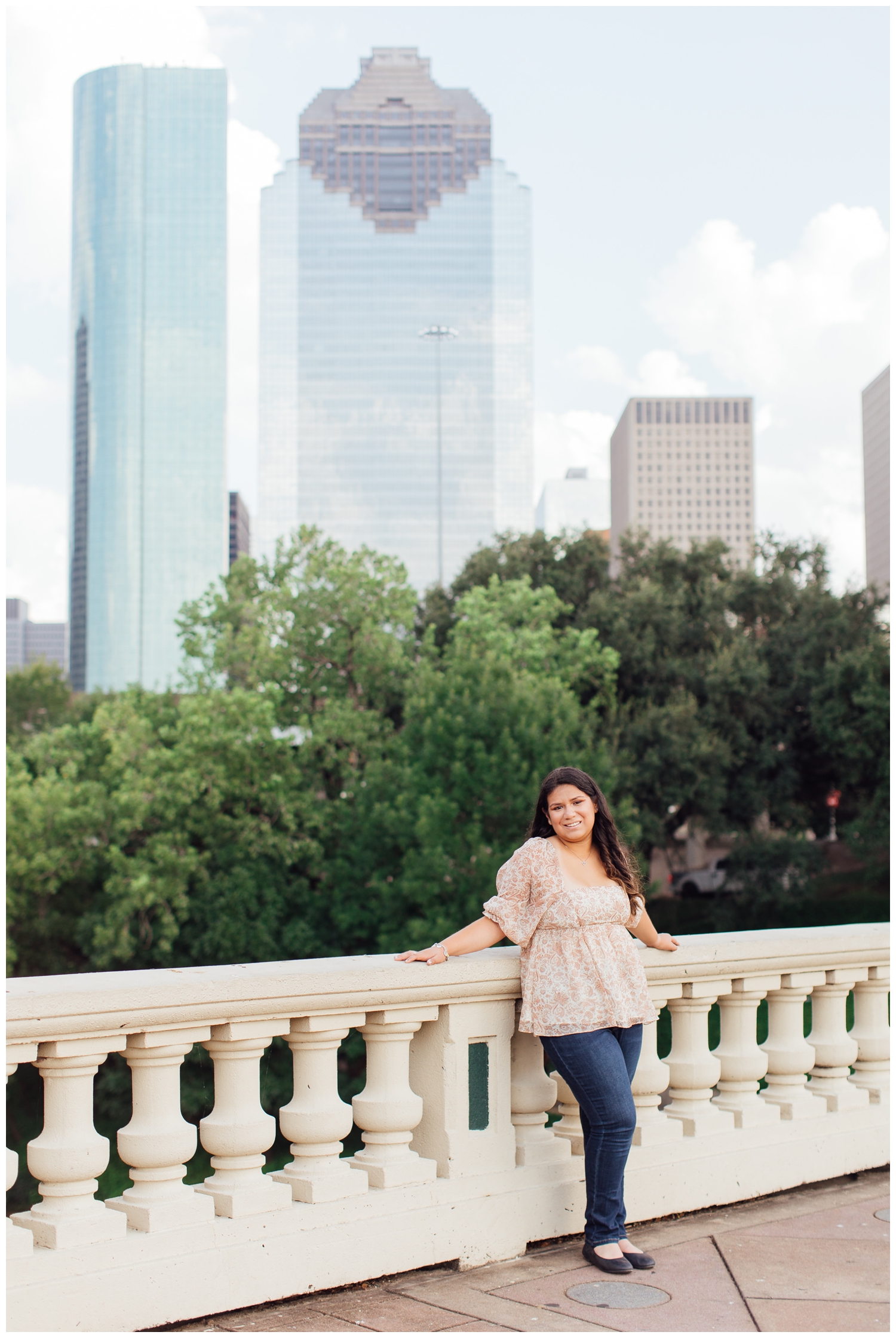 high school senior girl in floral top and denim jeans leaning on Sabine Street Bridge by Houston Skyline