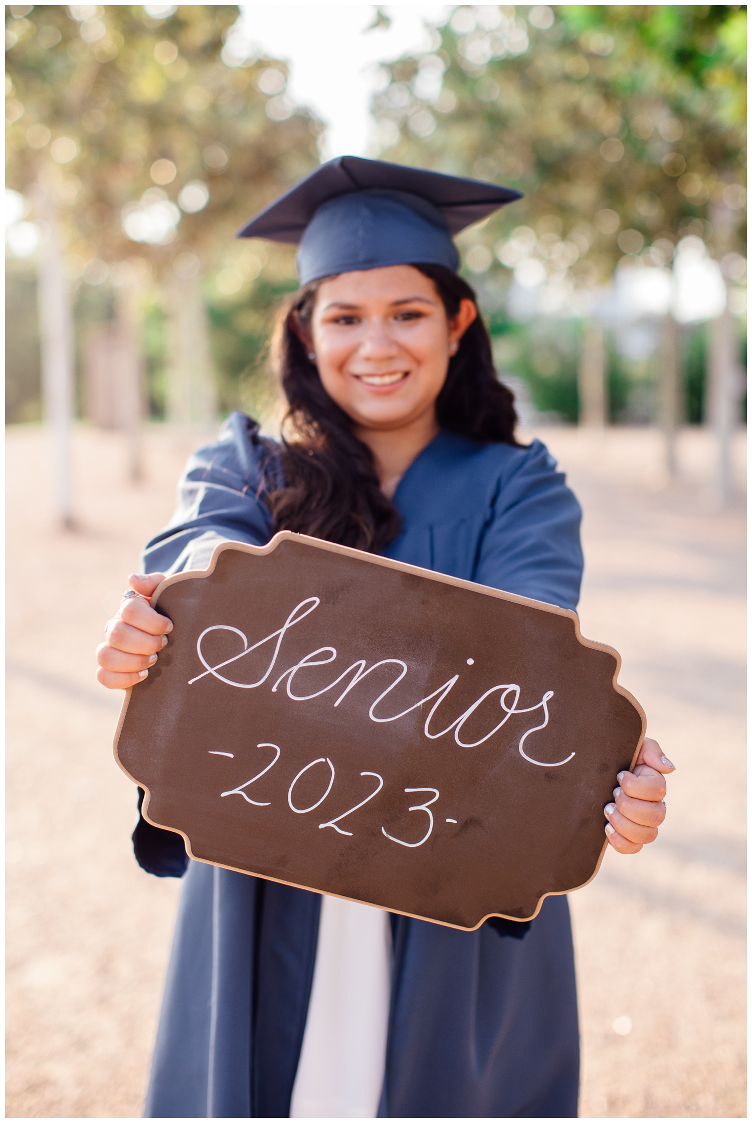 high school senior girl in blue cap and gown holding a senior sign outdoors at Sabine Street Houston