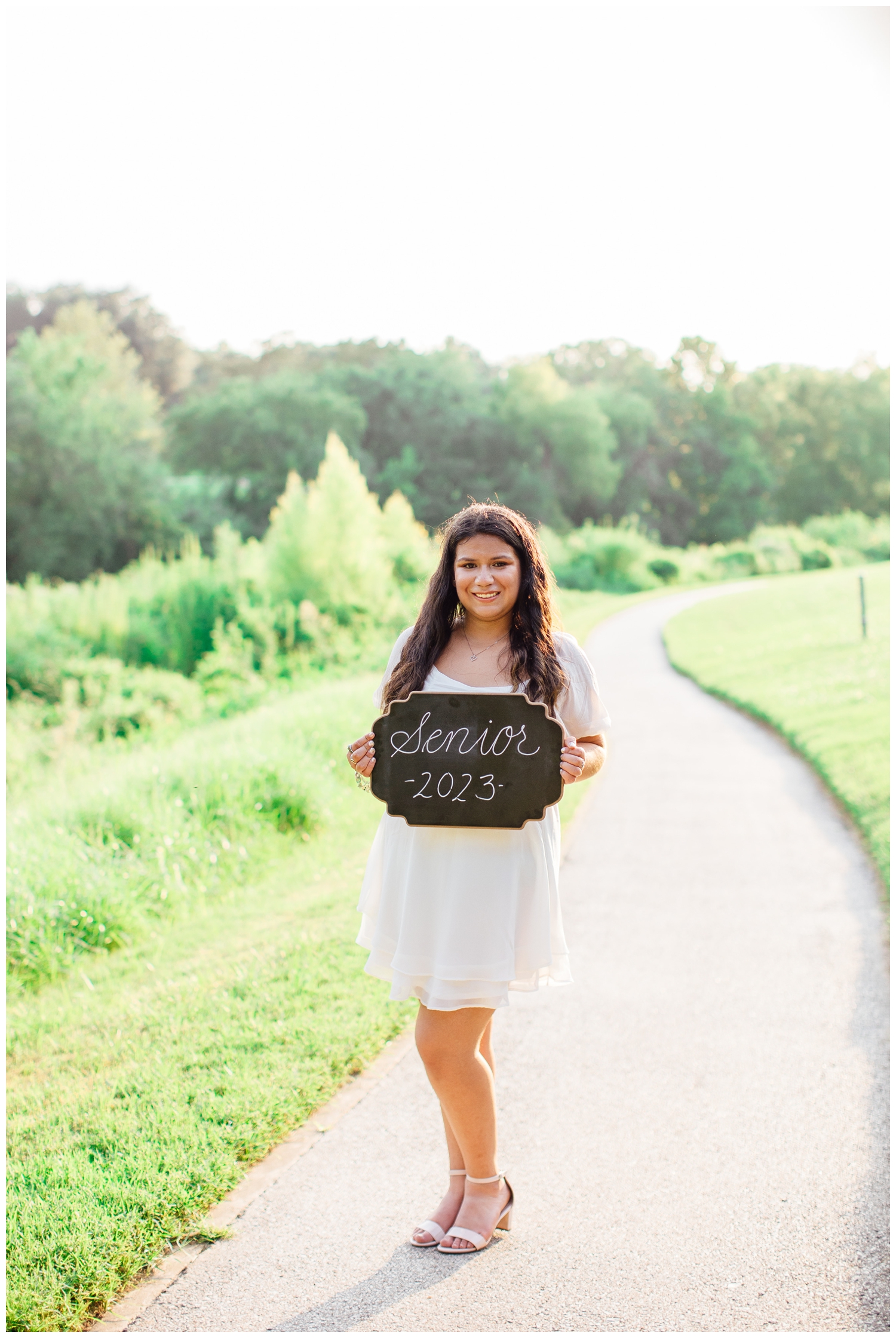 high school senior girl in white dress standing on pathway downtown Houston holding senior sign