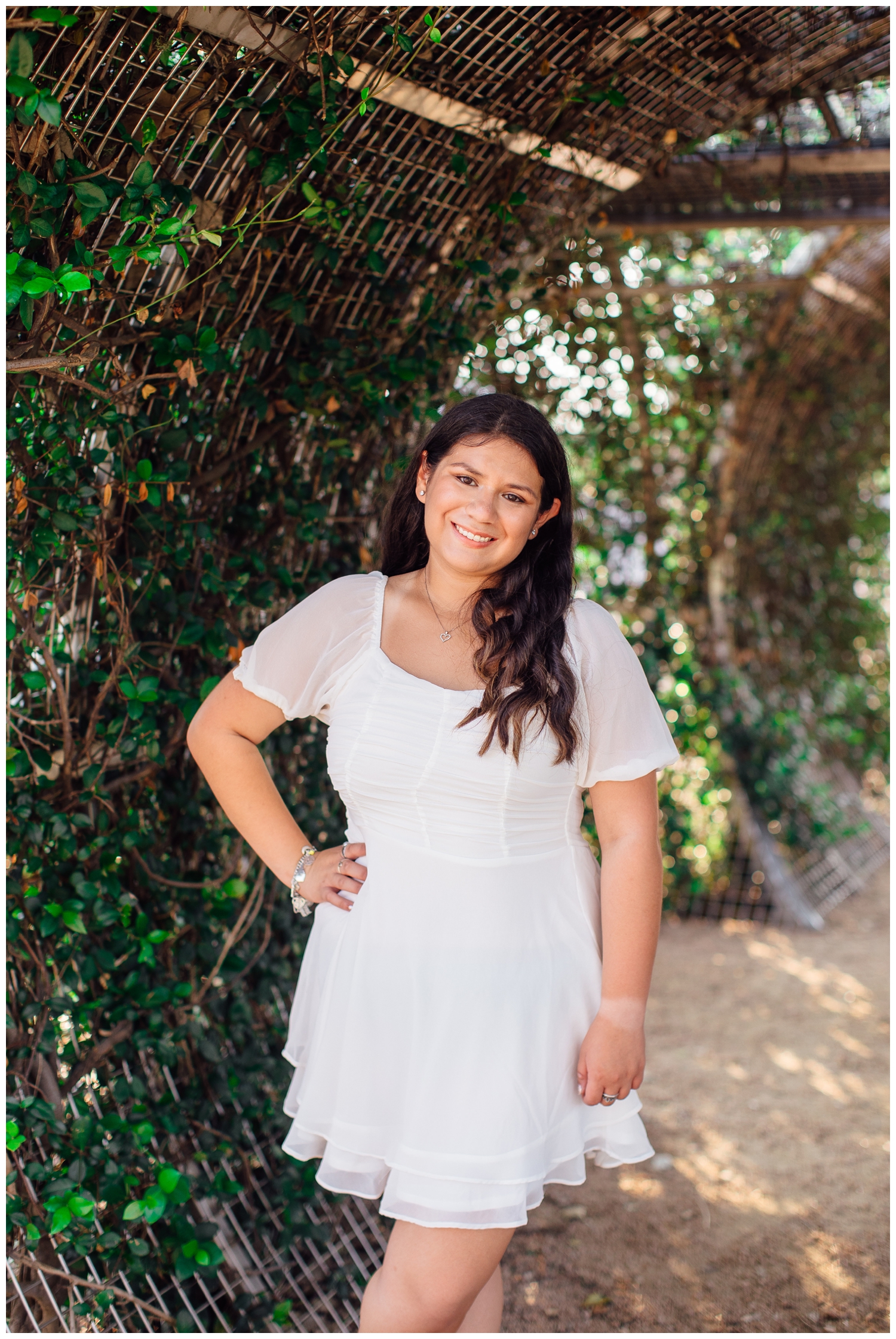 senior girl in white dress standing with hand on hip under vines for Senior Photos in Houston