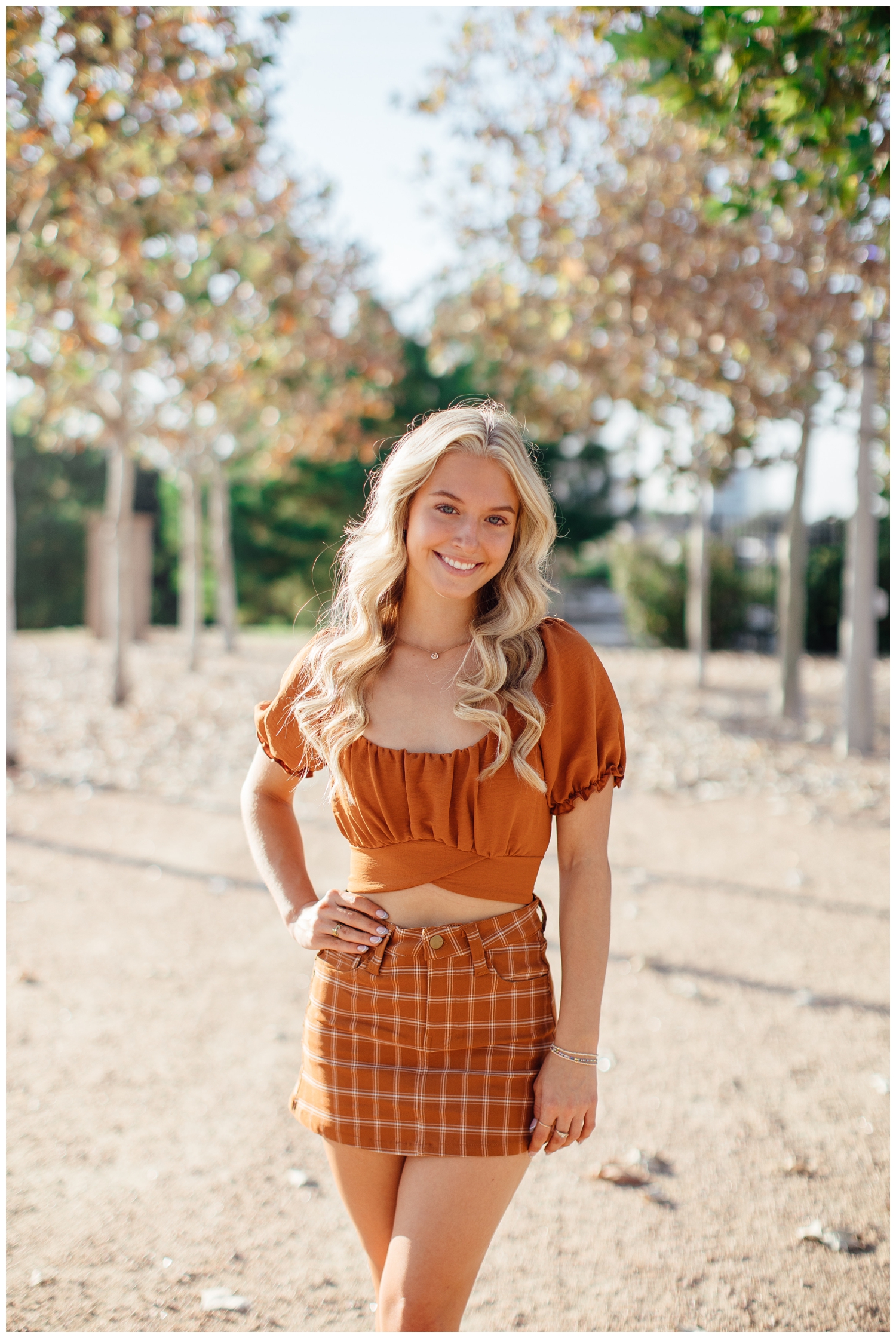 high school senior girl standing in downtown Houston tree line with hand in skirt pocket and smiling