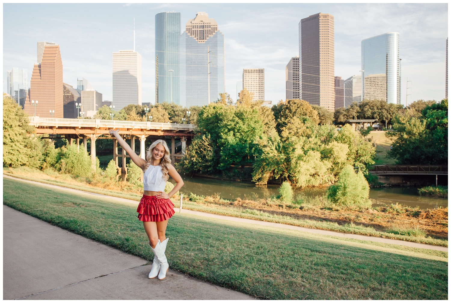 senior girl wearing white boots, white top and hot pink skirt with arm in air in front of downtown Houston skyline
