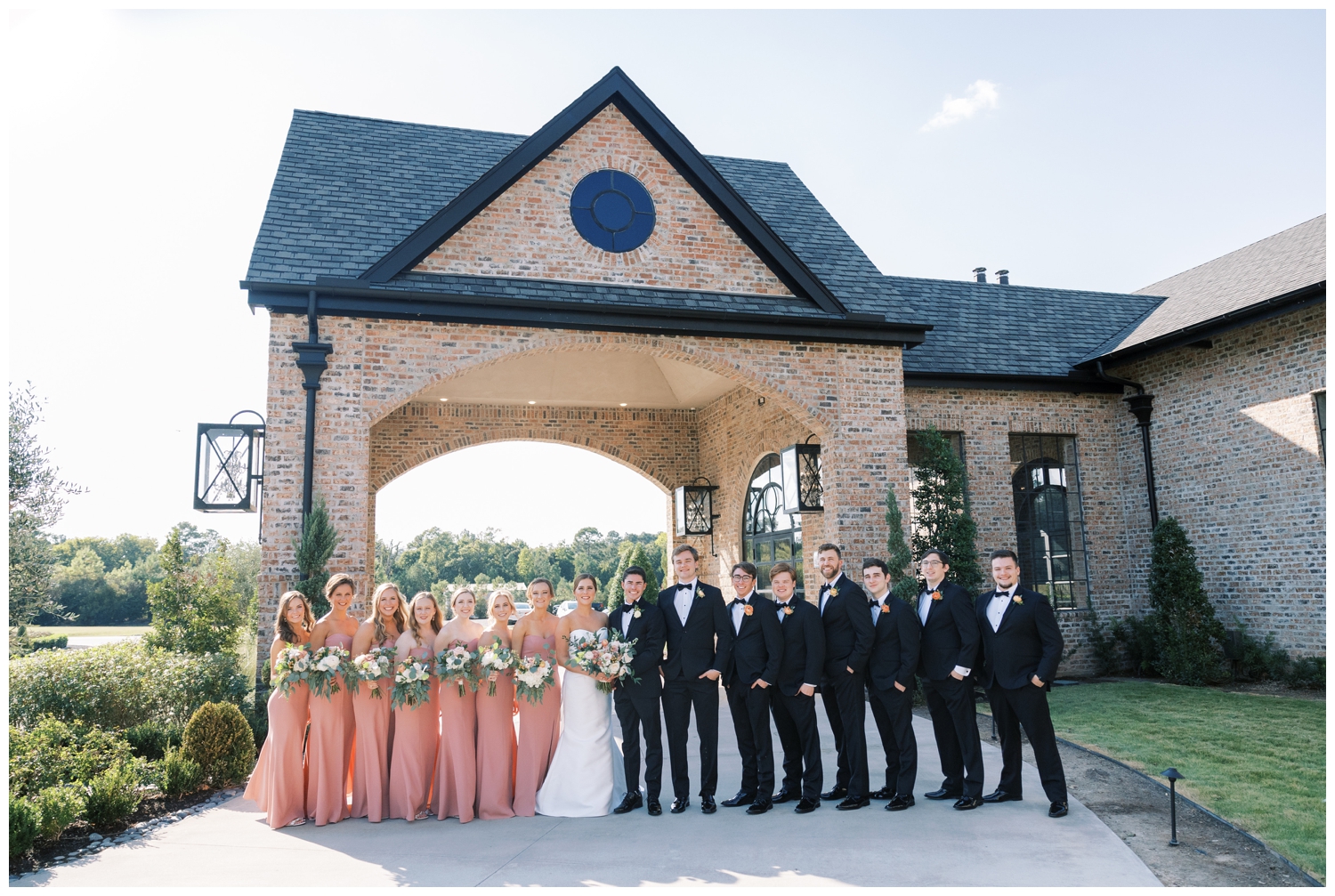 full wedding party in blush dresses and black tuxes standing in a line outside Iron Manor