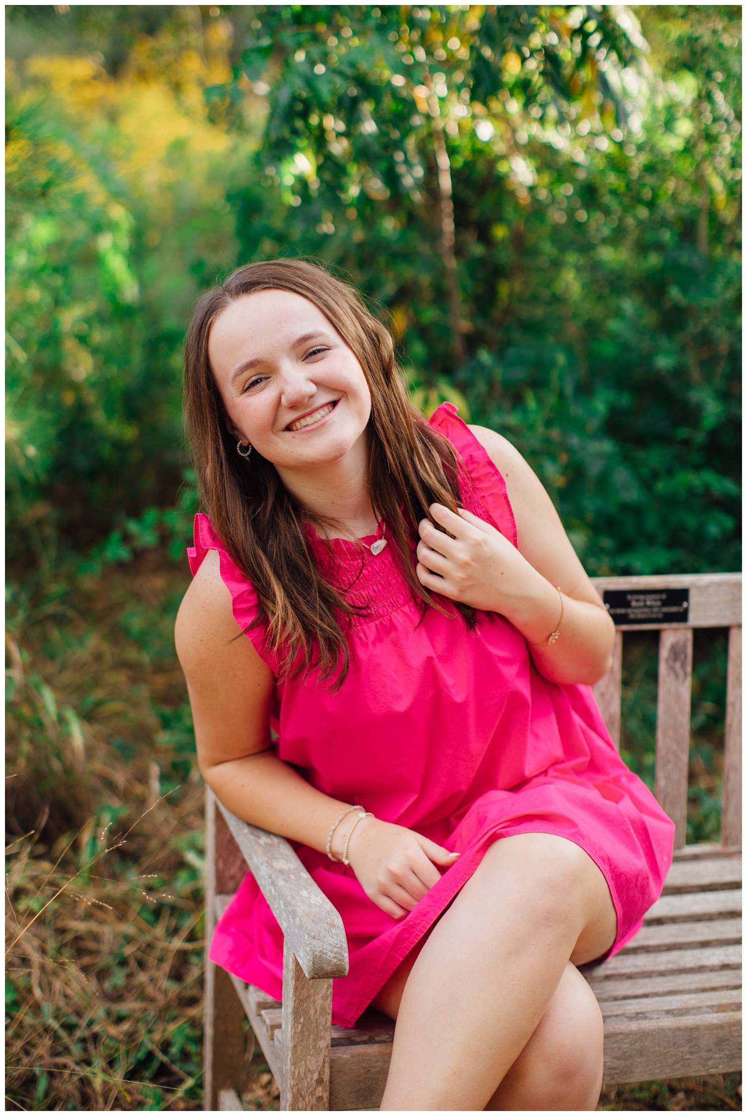 high school Houston senior in hot pink dress sitting on bench with hand in hair and smiling at Houston Arboretum