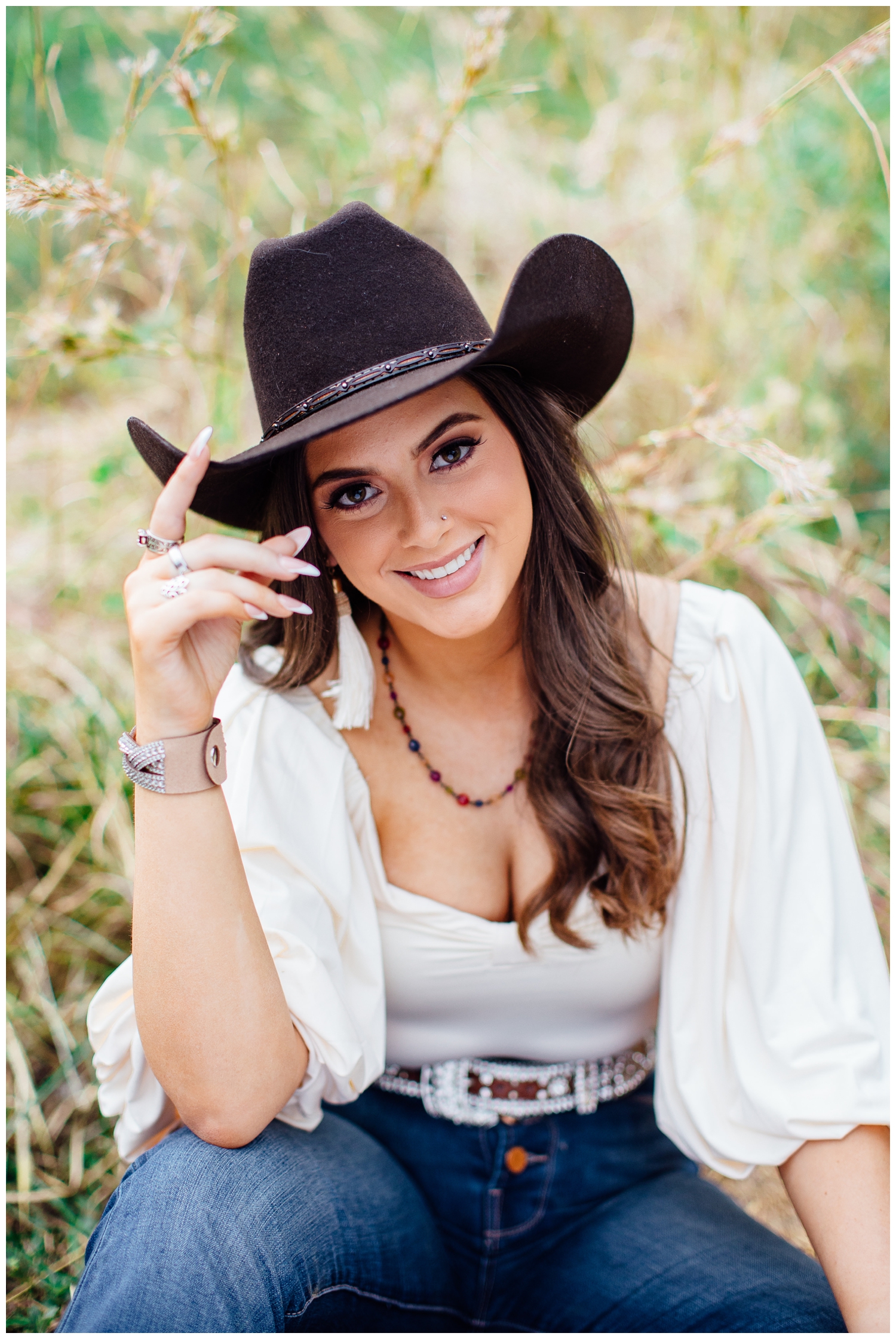 bold senior portraits in Houston Arboretum with girl sitting in field tipping black cowboy hat