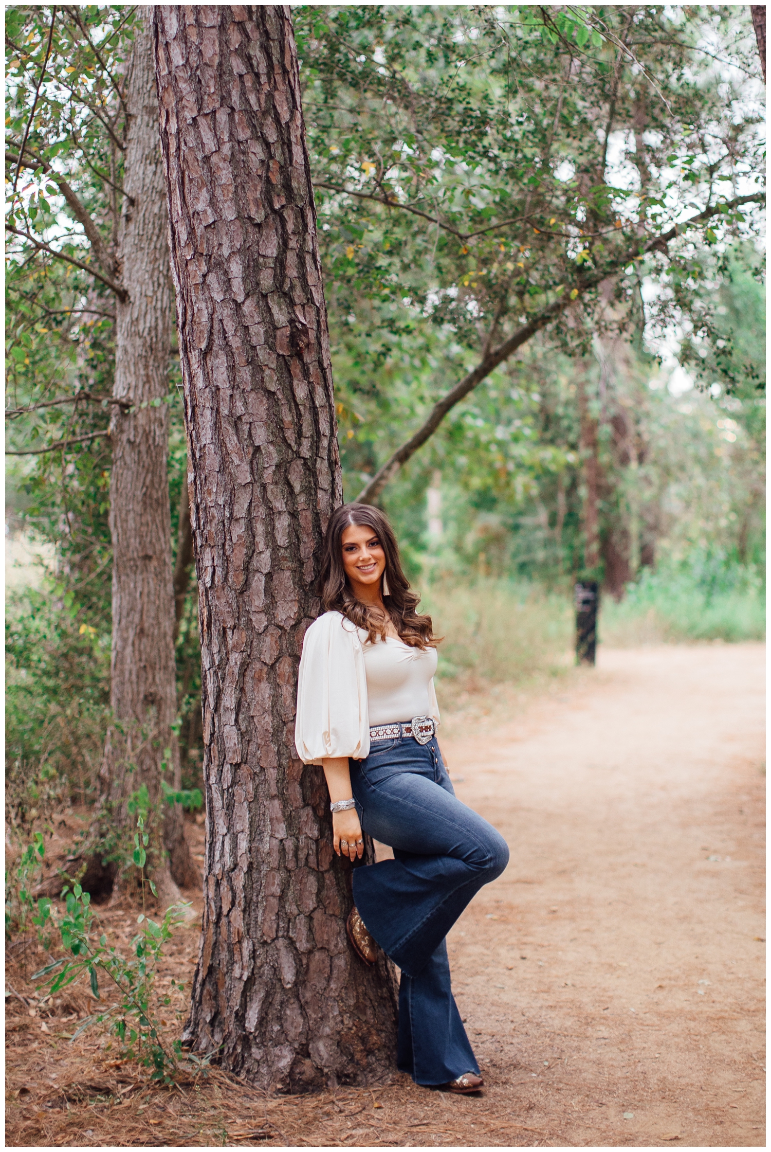 high school senior in jeans and cream shirt leaning against a tree outdoors at Houston Arboretum