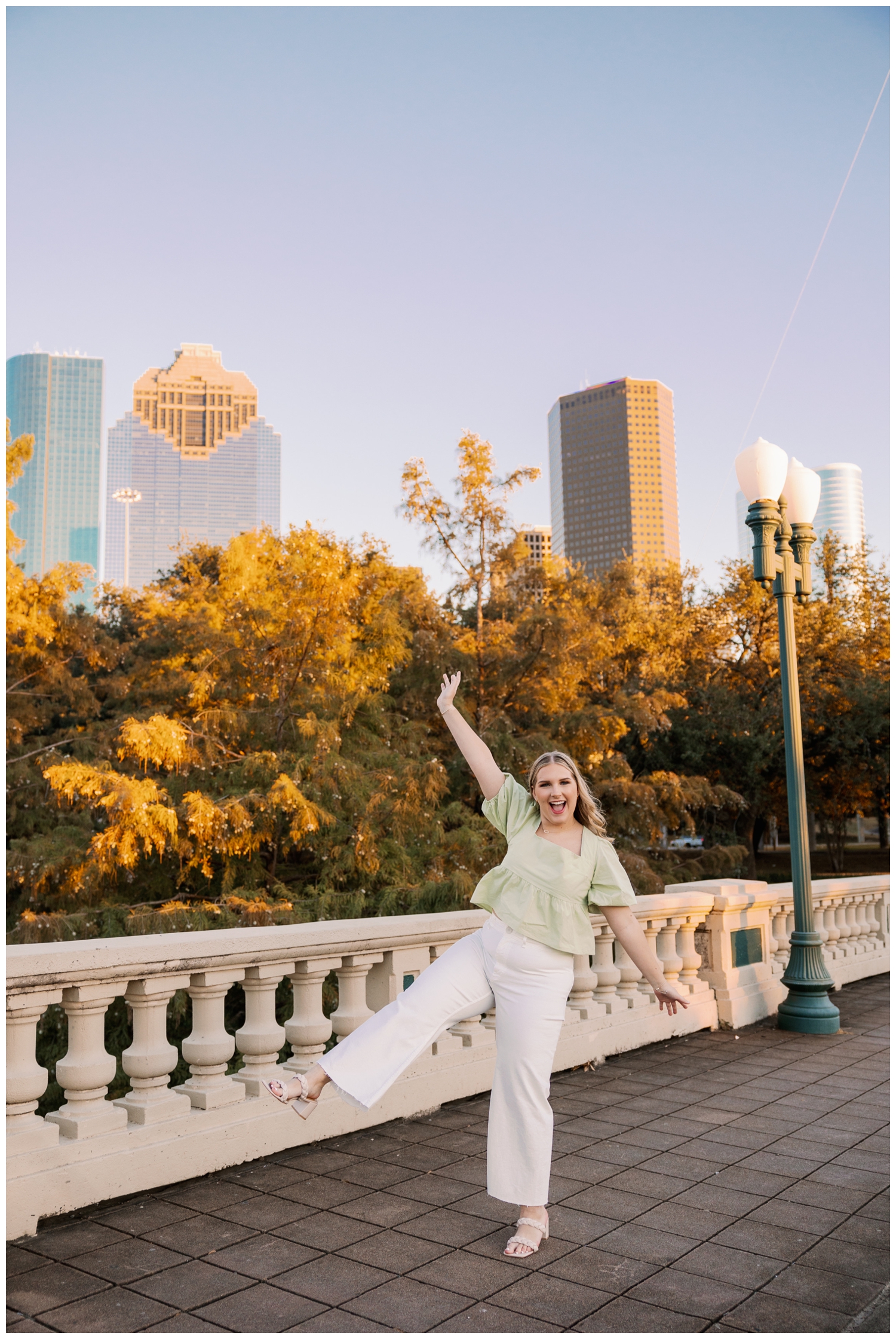 girl in white jeans and green blouse kicking legs on Sabine Street bridge in font of Houston skyline