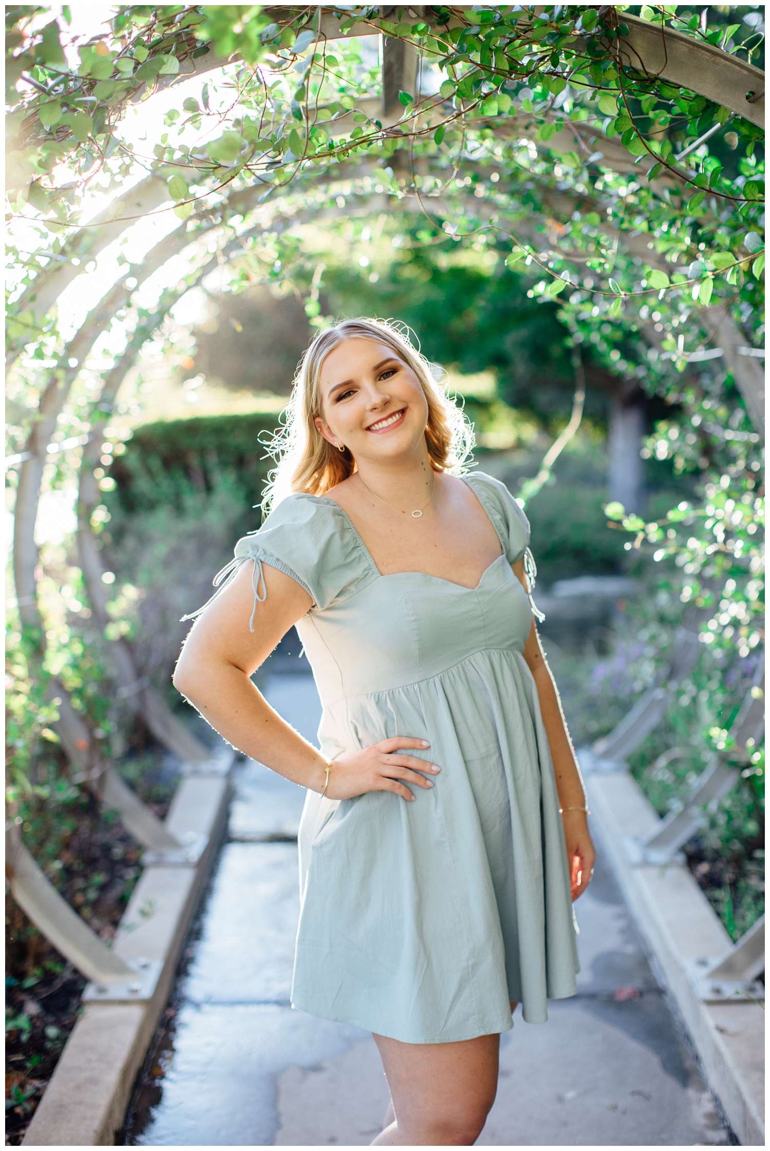 girl in sage dress standing hand on hip under garden arch for cute senior pictures in Houston