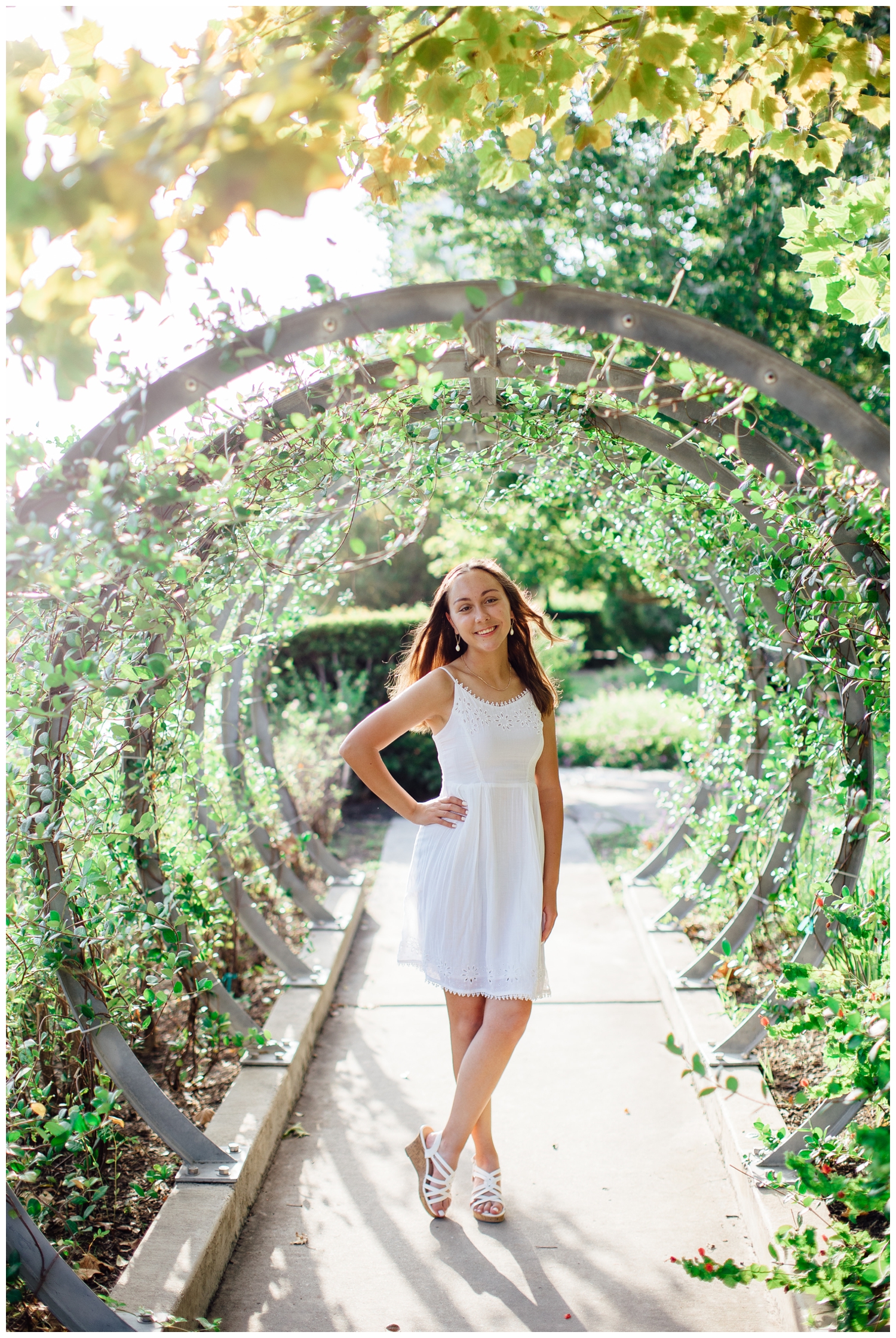 senior girl in white sundress standing under silver arches with greenery for downtown Houston senior pictures