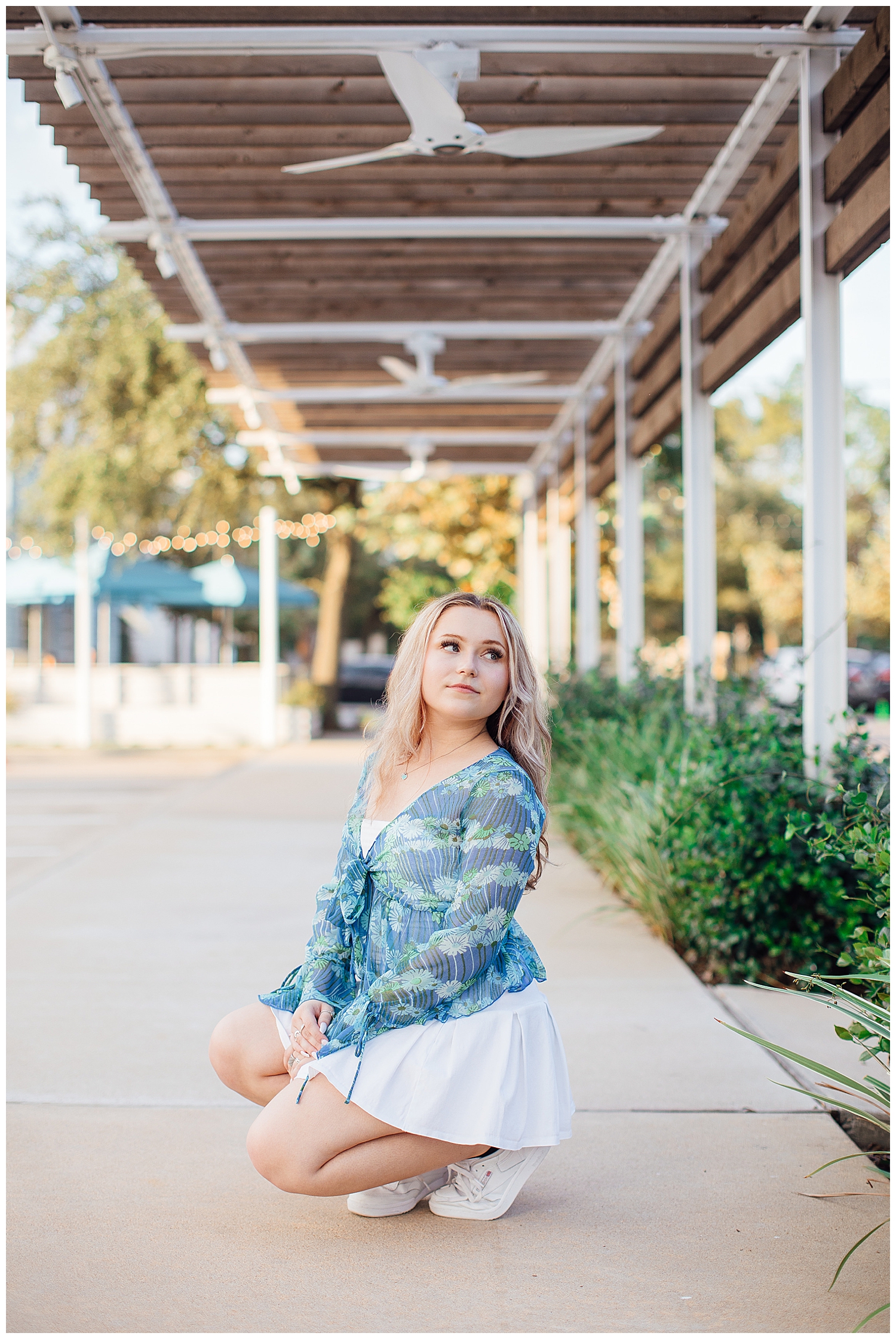 high school senior standing under walkway at Uptown Park Houston wearing white skirt blue shirt