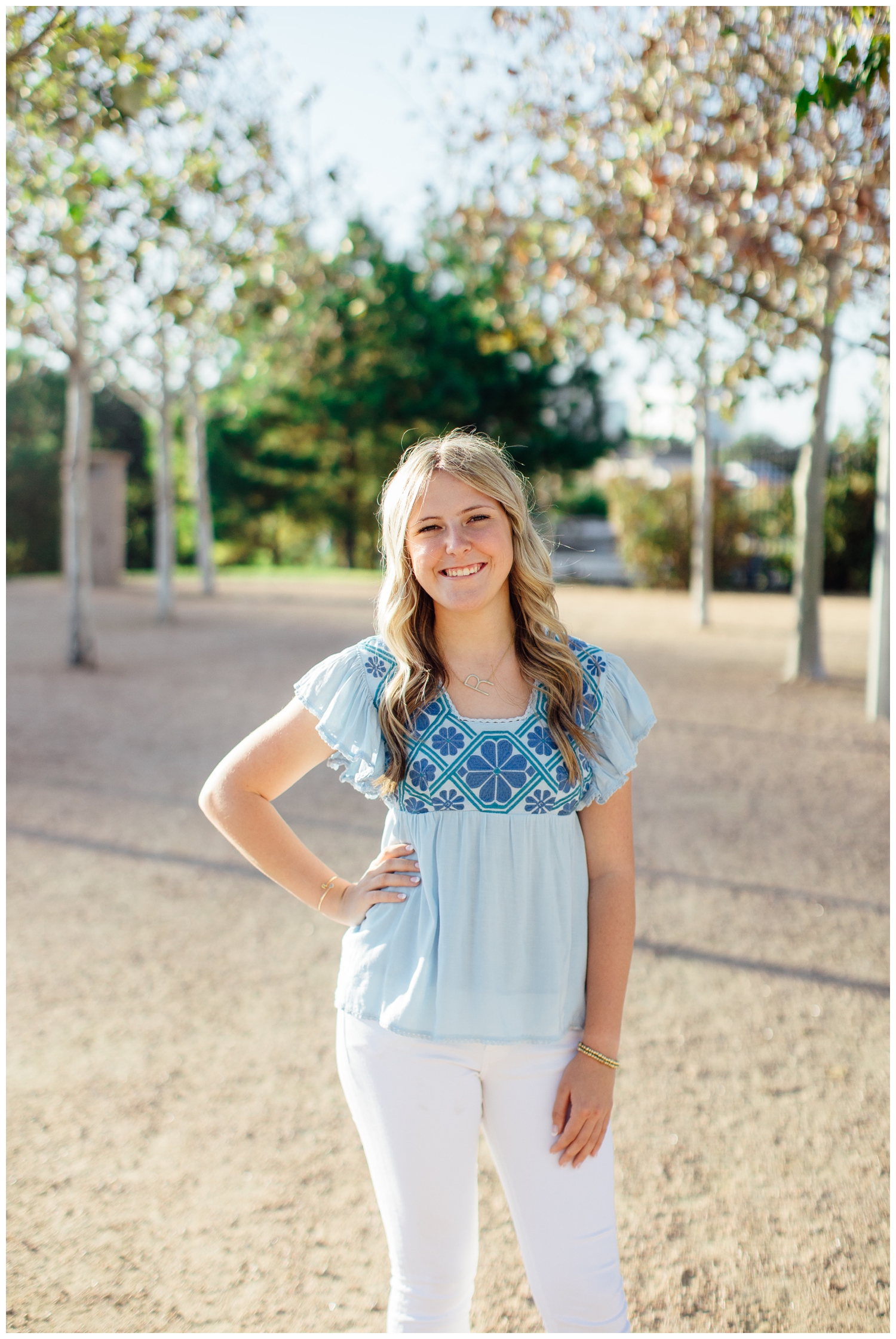 high school senior girl in light blue blouse white pants standing between trees