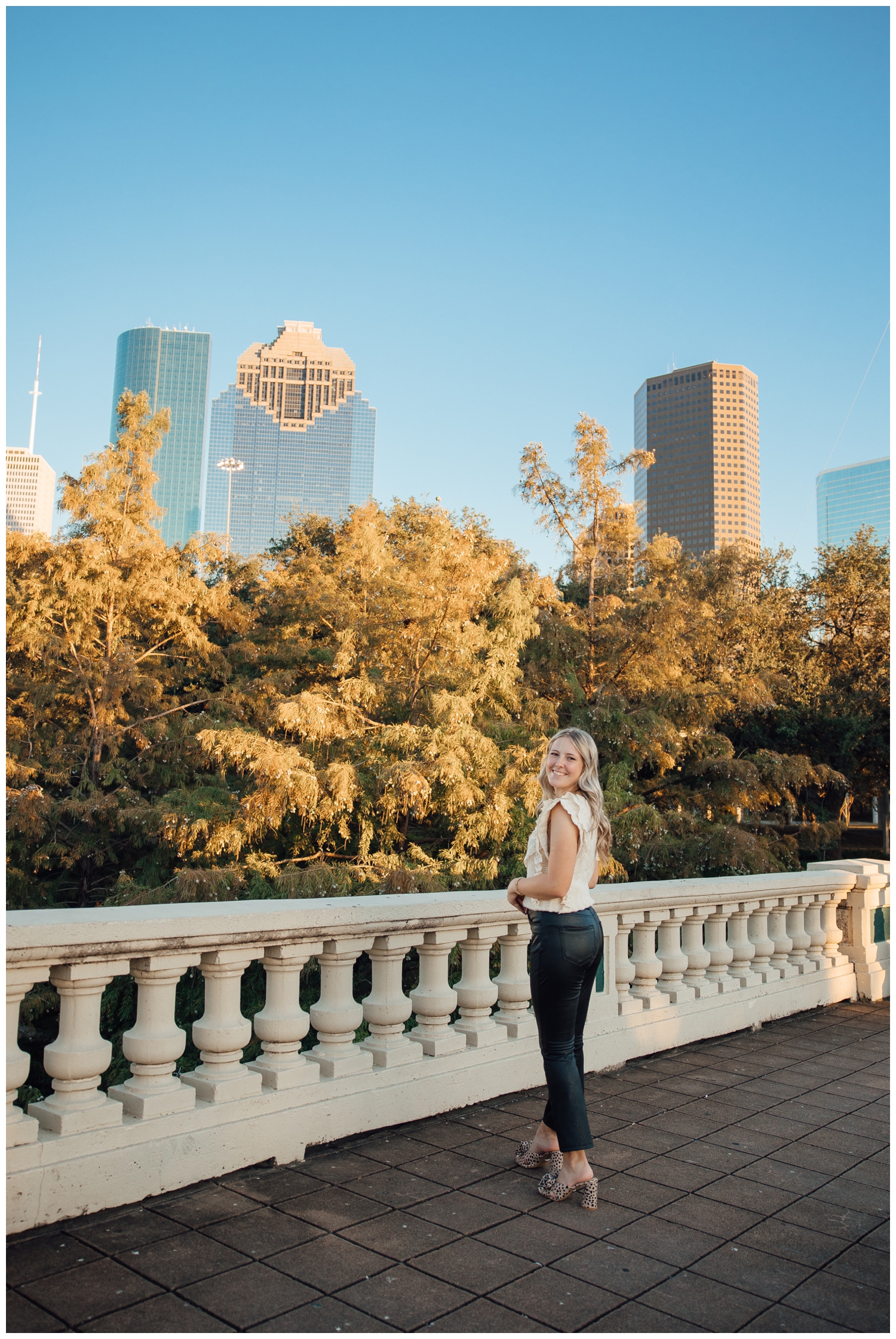 Outdoor Houston senior photography with girl in black leather pants cream shirt standing in front of Houston skyline