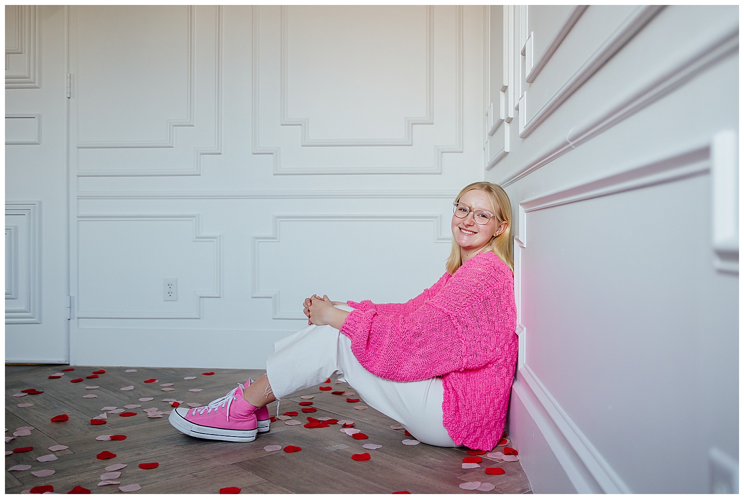 high school girl in hot pink shirt, converse and white pants sitting for Valentine's Day Senior Photoshoot