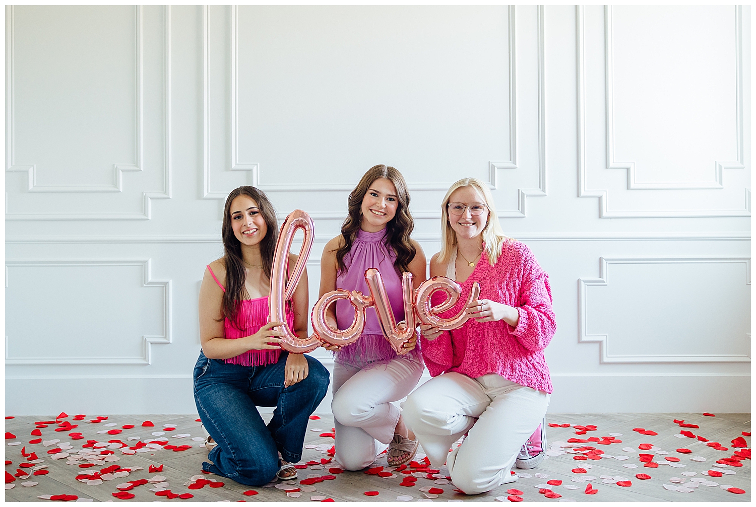 three senior girls kneeling on rose petals holding pink love balloon for Valentine's Day Senior Photoshoot