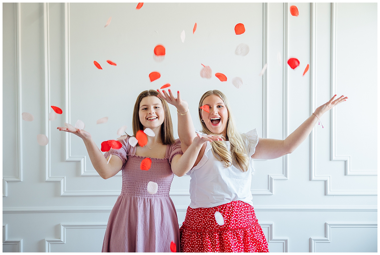 two senior girls tossing rose petals in front of white studio wall