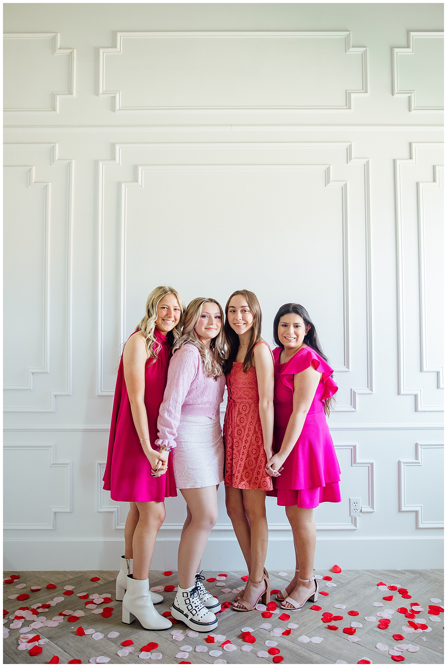 four girls in pink, red and white dresses holding hands in front of white wall smiling