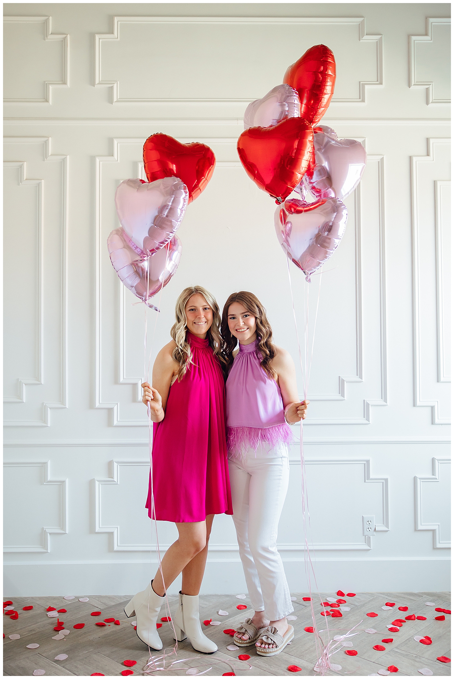 two high school senior girls standing in front of white studio wall for Valentine's Day Senior Photoshoot holding heart balloons