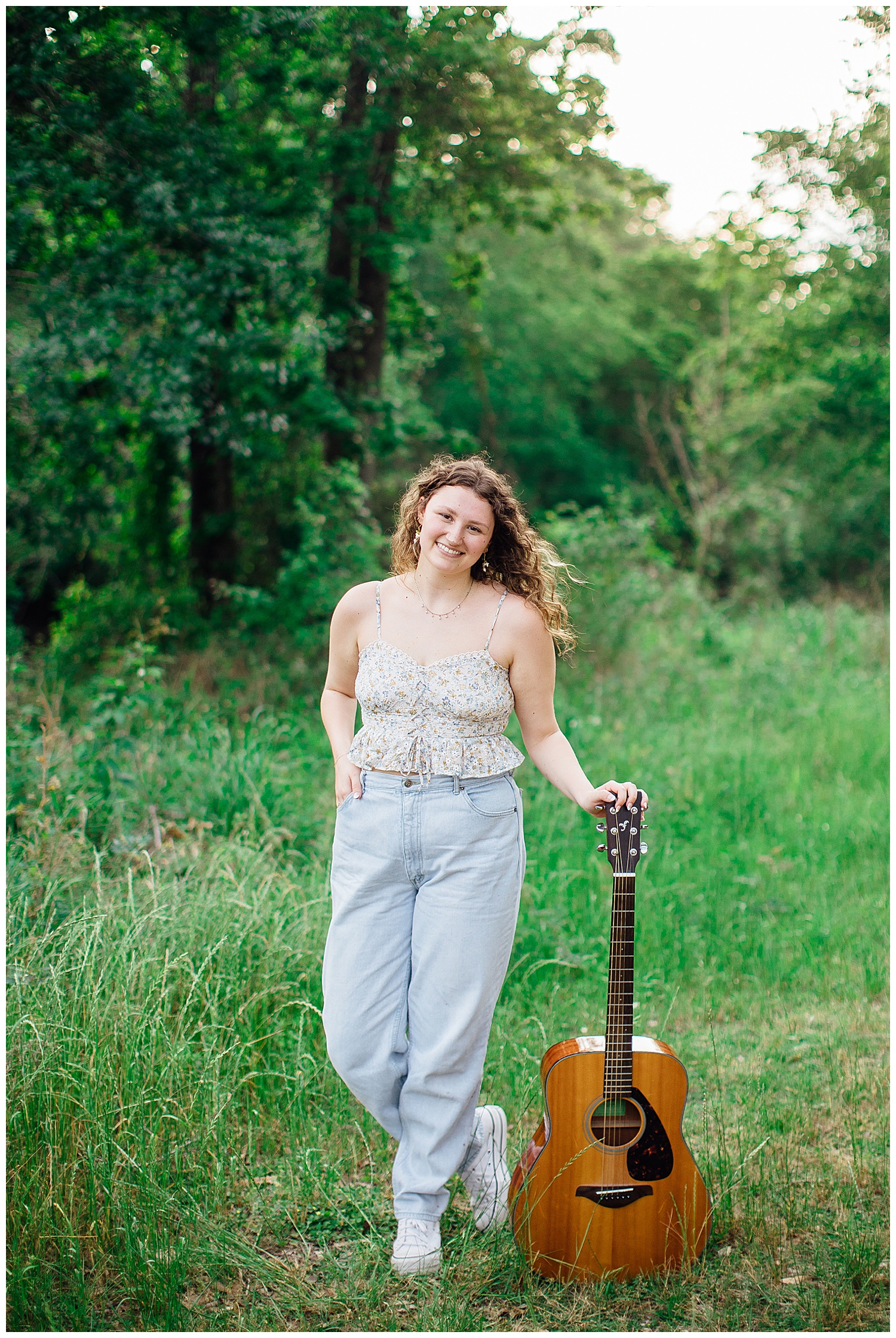high school senior girl standing in jeans and cream top in a field with guitar