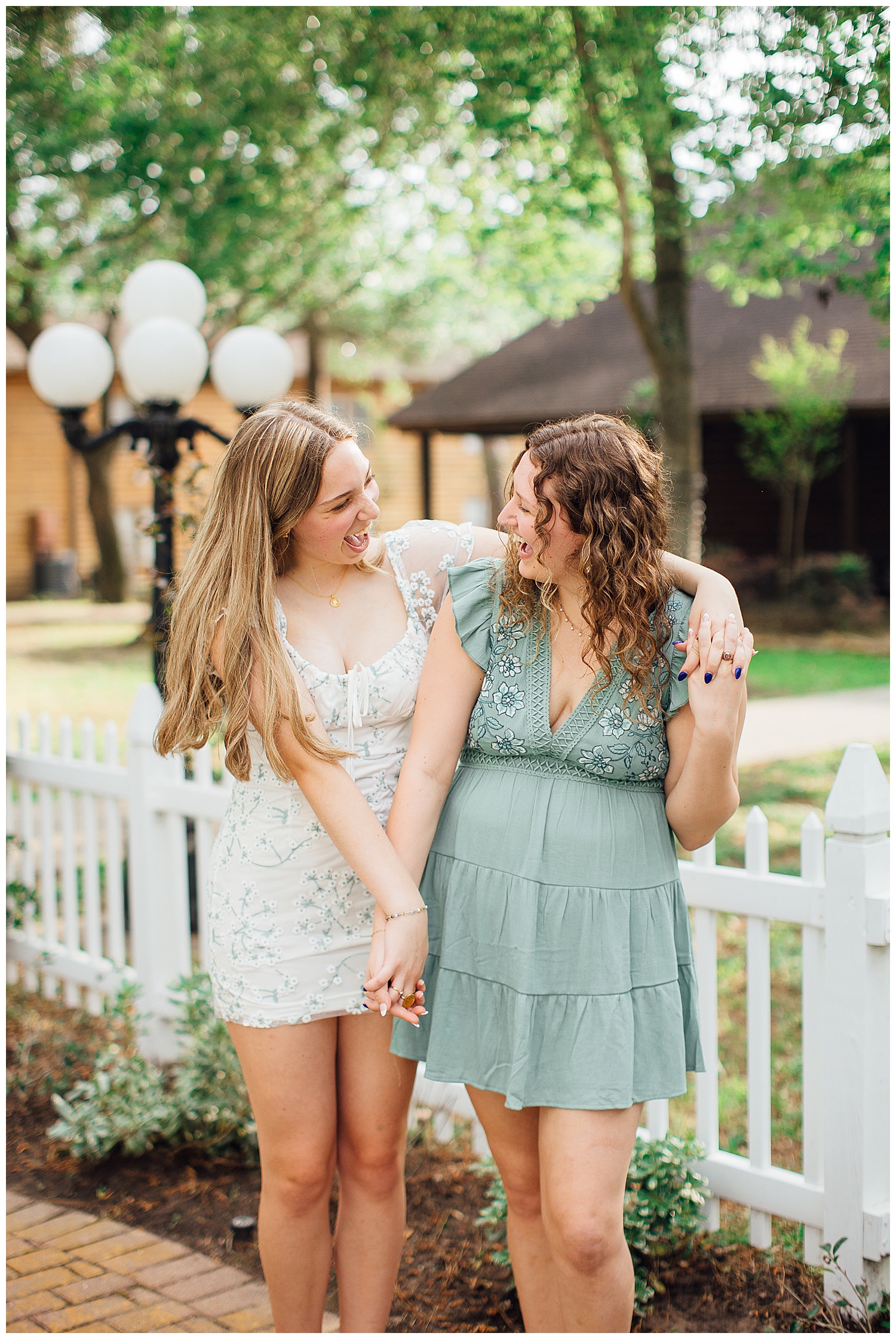 two best friends smiling and laughing outdoors near a white picket fence for best friends senior pictures