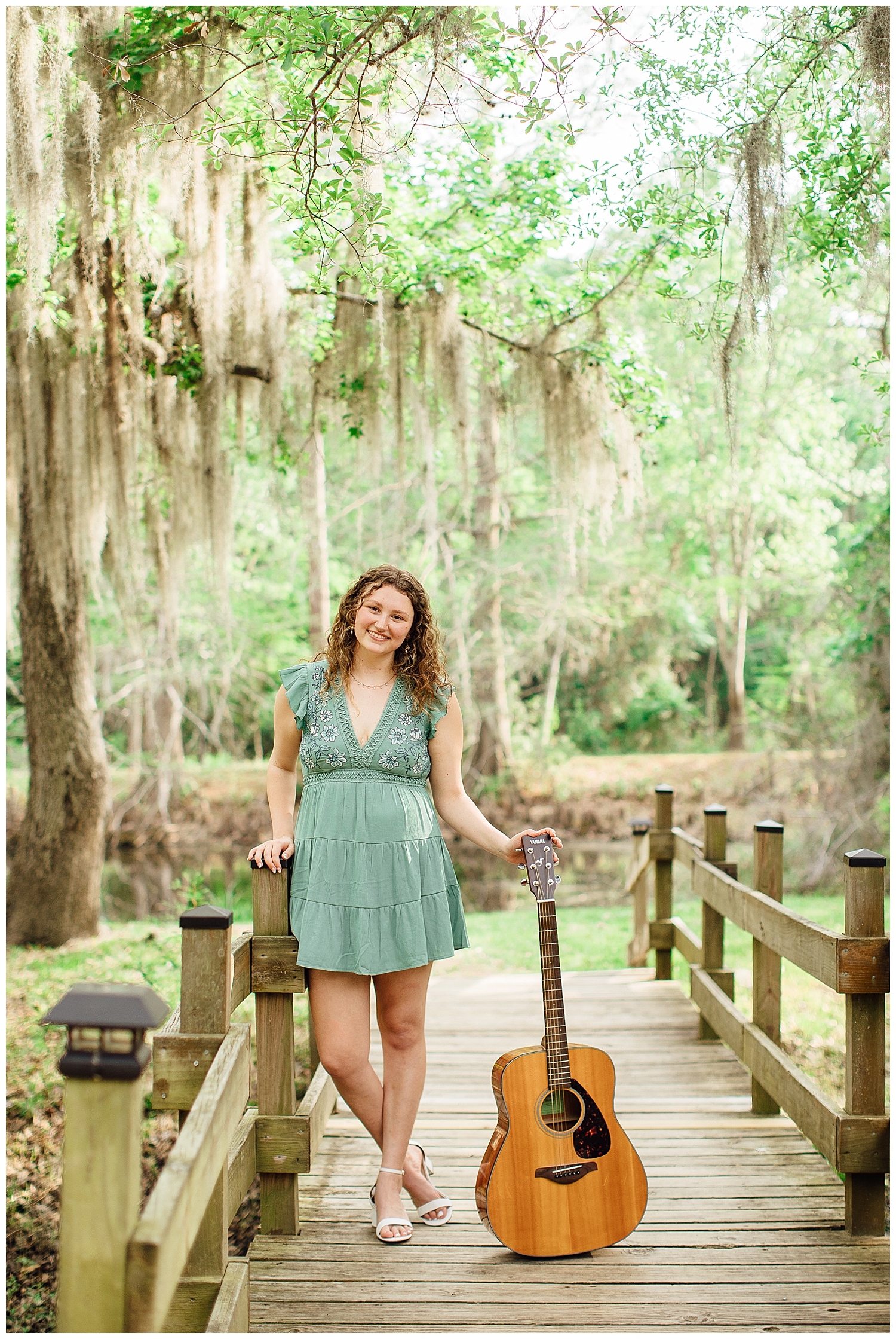 senior girl in green dress standing under mossy trees on a bridge at Cy Hope with a guitar