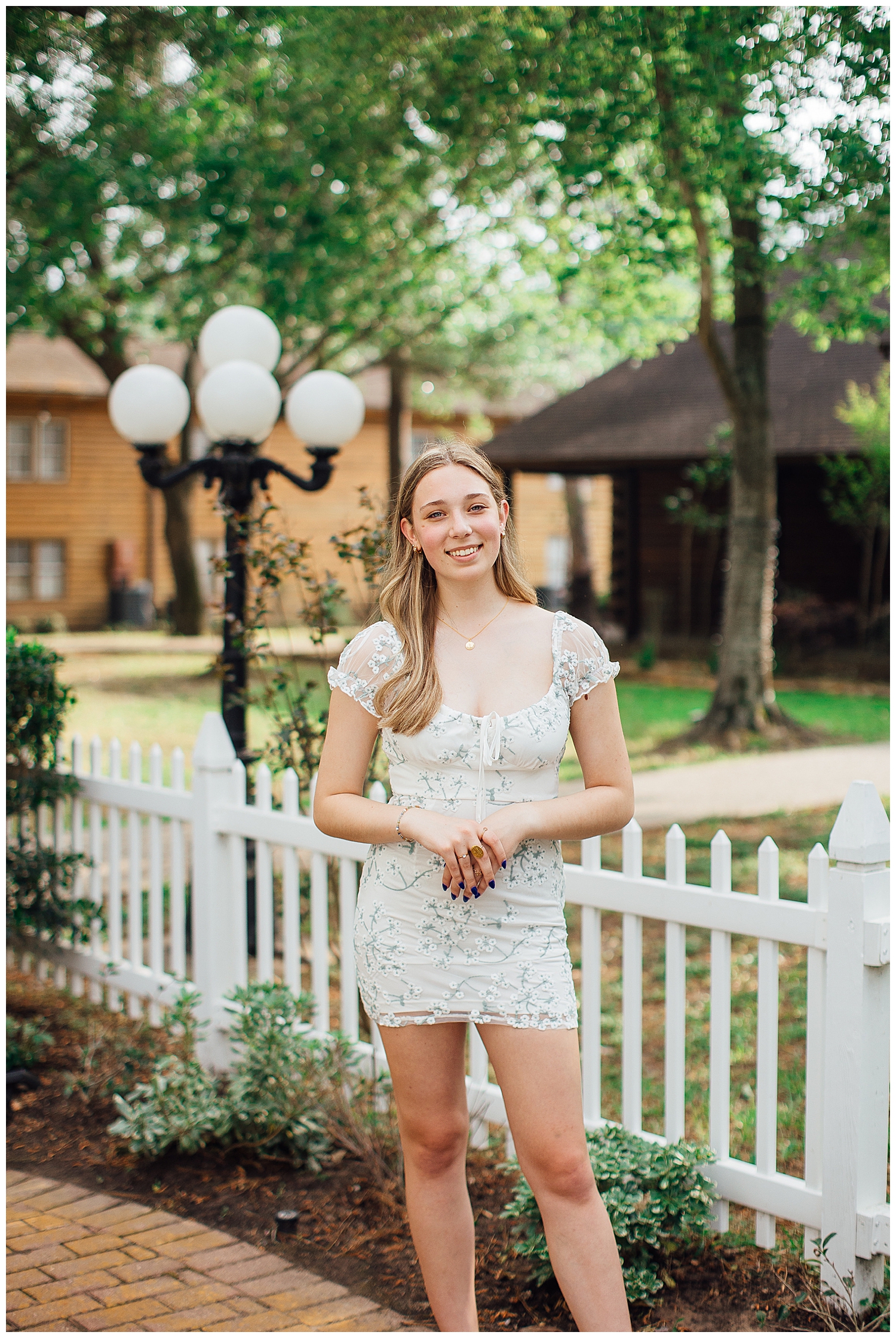 senior girl standing in cream dress in front of white picket fence with hands crossed